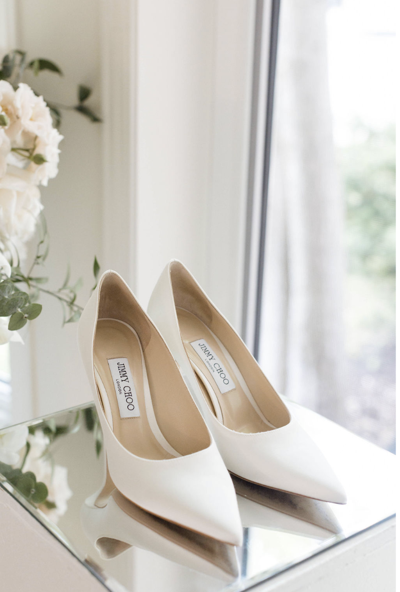 Une paire de chaussures à talons blancs de la marque Jimmy Choo, posées sur un miroir près d'une fenêtre, avec des fleurs blanches en arrière-plan.