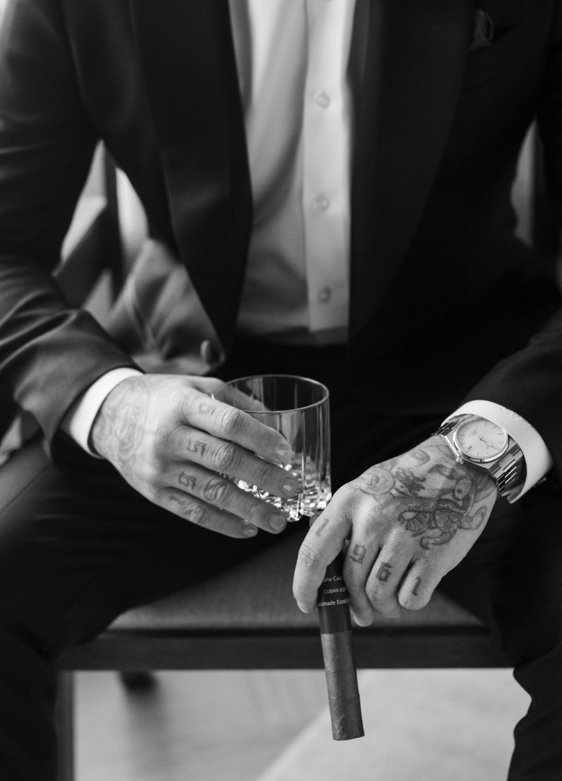 Homme assis portant un costume noir, une montre et plusieurs tatouages, tenant un verre à whisky et une cigare.
