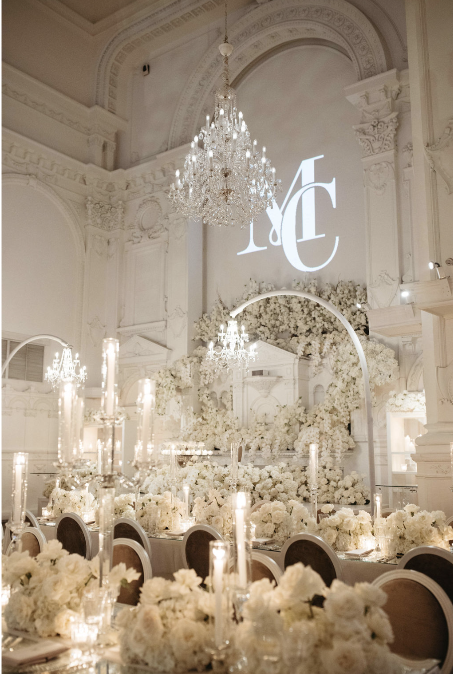 Salle de mariage élégante à la Maison Principale à Montréal avec décoration blanche, chandeliers, fleurs, et une projection de monogramme sur le mur.