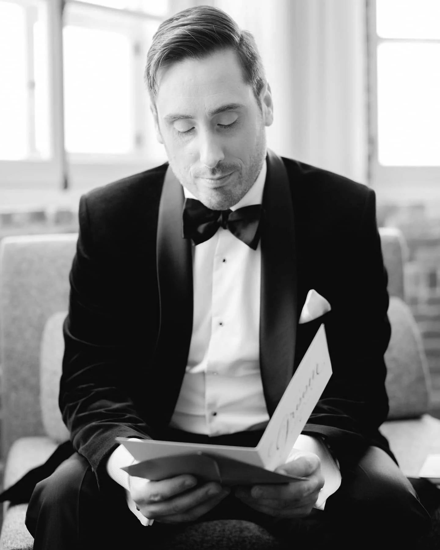 Futur marié en smoking avec nœud papillon, regardant un livre, assis dans une pièce lumineuse