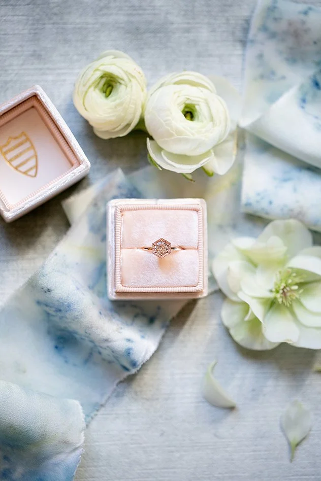 Une bague en or avec un diamant au centre, placée dans une boîte en velours rose, entourée de fleurs blanches et de tissus doux.