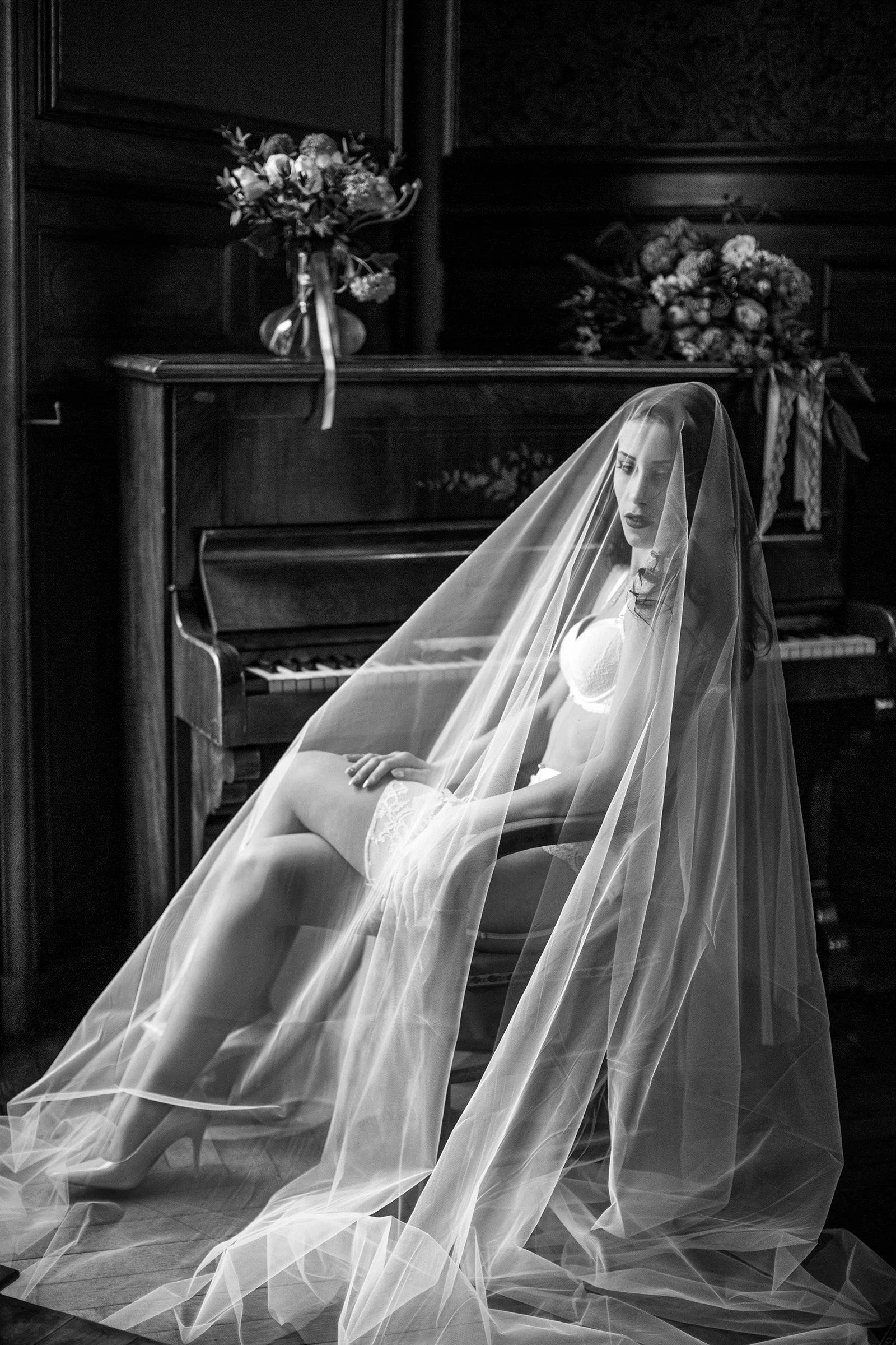 Femme en lingerie blanche et voile de mariée assise devant un piano en bois avec des bouquets de fleurs sur le meuble, photo en noir et blanc.