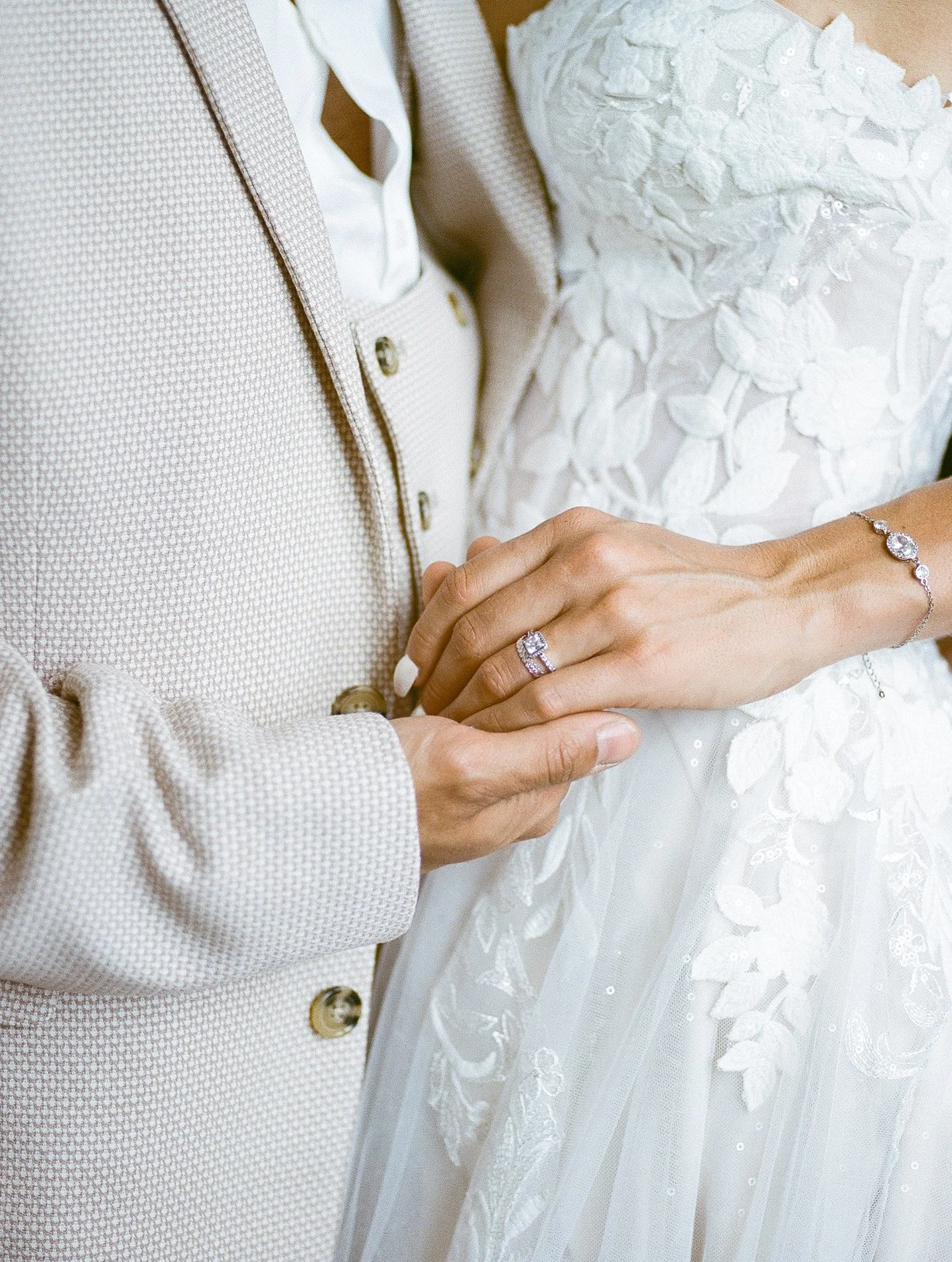 Zoom sur deux personnes en robe de mariée et costume, mettant en valeur une alliance et un bracelet sur la main de la mariée.