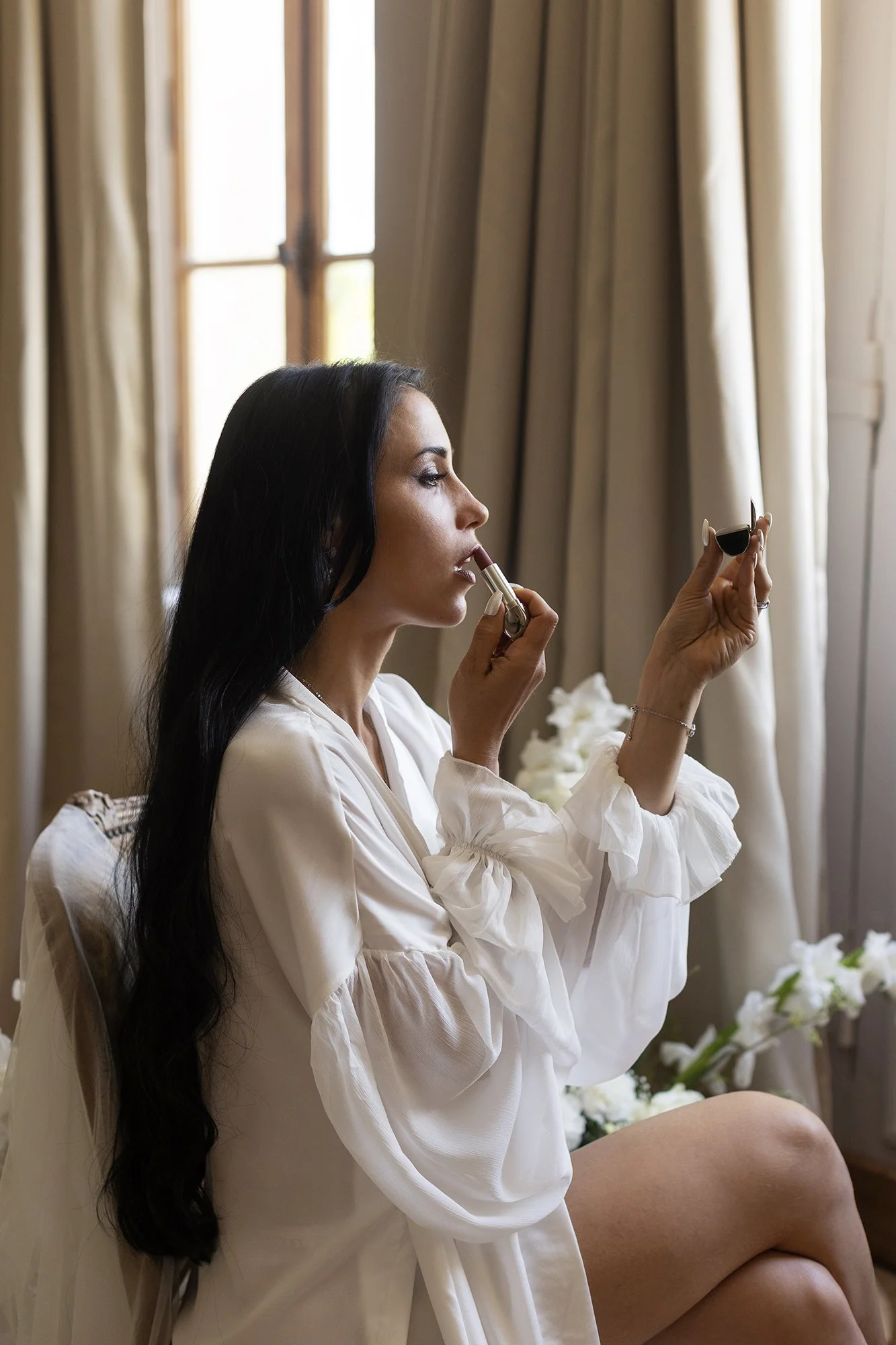 Une future mariée au château Fengari aux cheveux longs et noirs se maquille les lèvres en utilisant un rouge à lèvres devant un miroir, dans une pièce lumineuse avec des rideaux beiges et des fleurs blanches sur le côté.