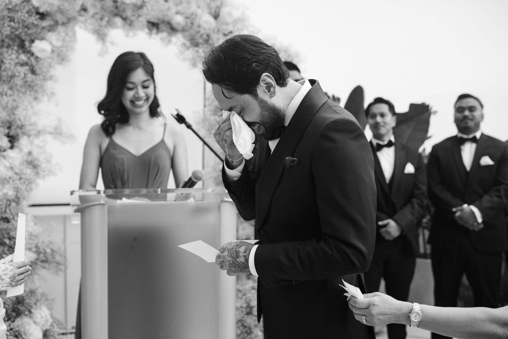 Un homme en veste de costume noir, visiblement ému, essuie ses yeux lors d'une cérémonie de mariage. Une femme à l'arrière sourit, et plusieurs hommes en costard homologue en arrière-plan. L'événement se déroule à l'extérieur.