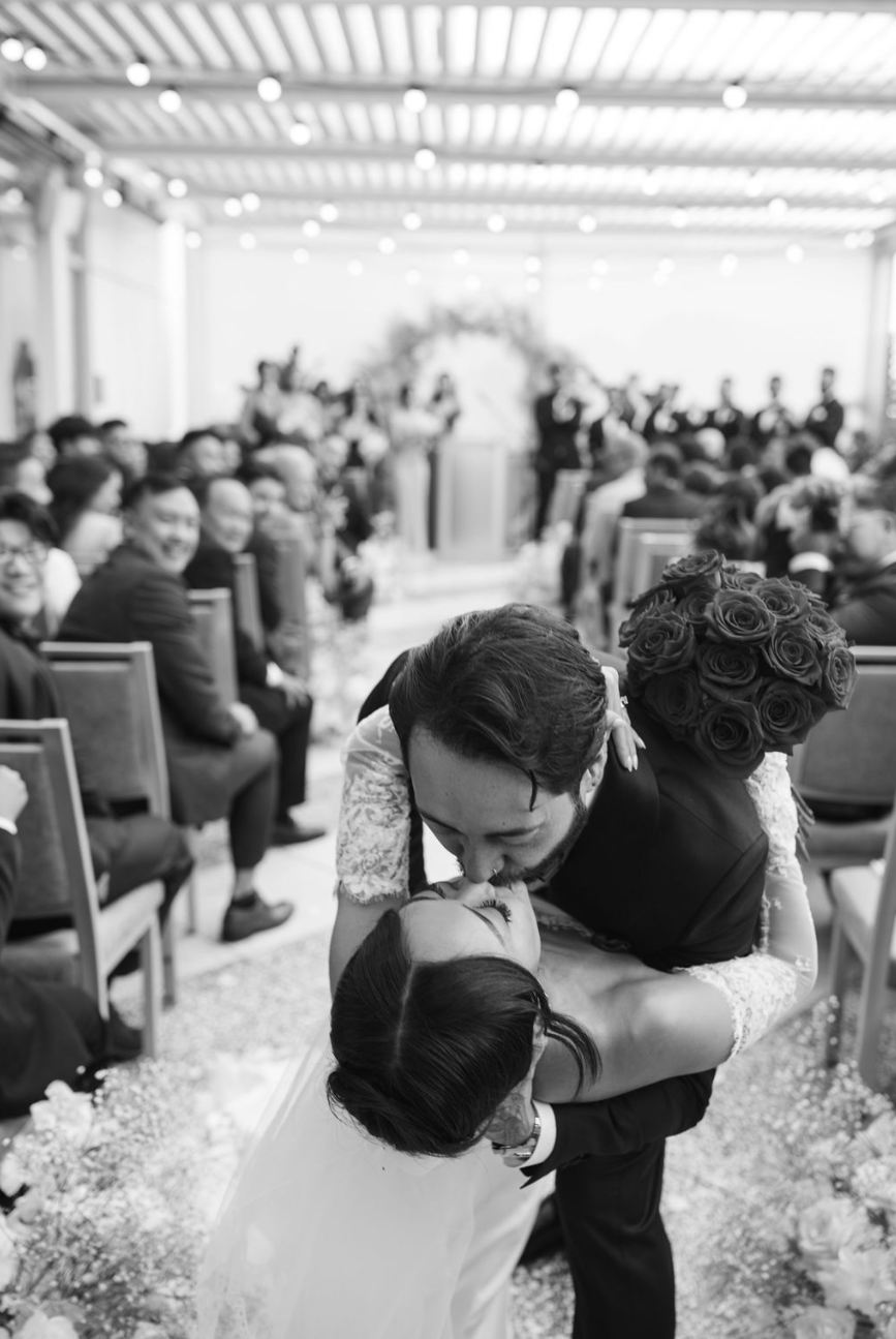 Un couple de mariés s'embrassant lors de leur mariage en intérieur, entouré de nombreux invités assis et regardant la scène.