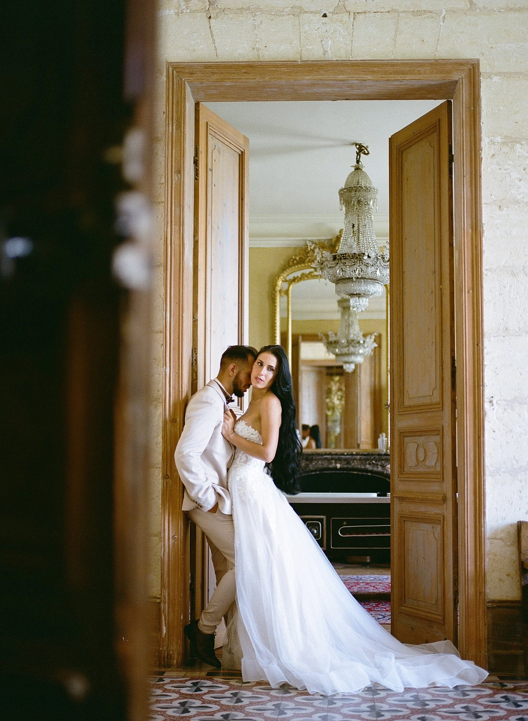 Un couple en habits de mariage posant au chateau Fengari en France avec des lustres en cristal et des miroirs dorés.