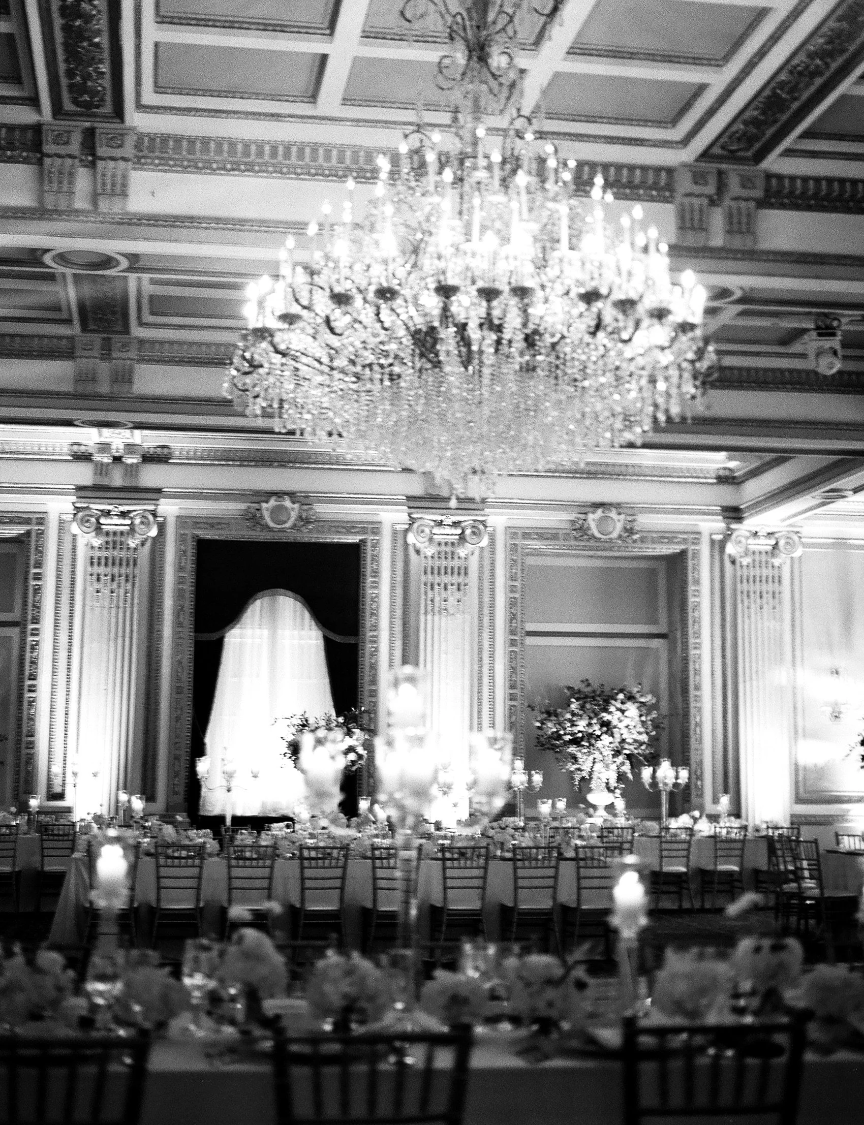 Une salle de réception élégante avec un grand chandelier suspendu au plafond, des murs ornés de moulures, des vases de fleurs, des tables dressées pour un dîner, et des chandelles.