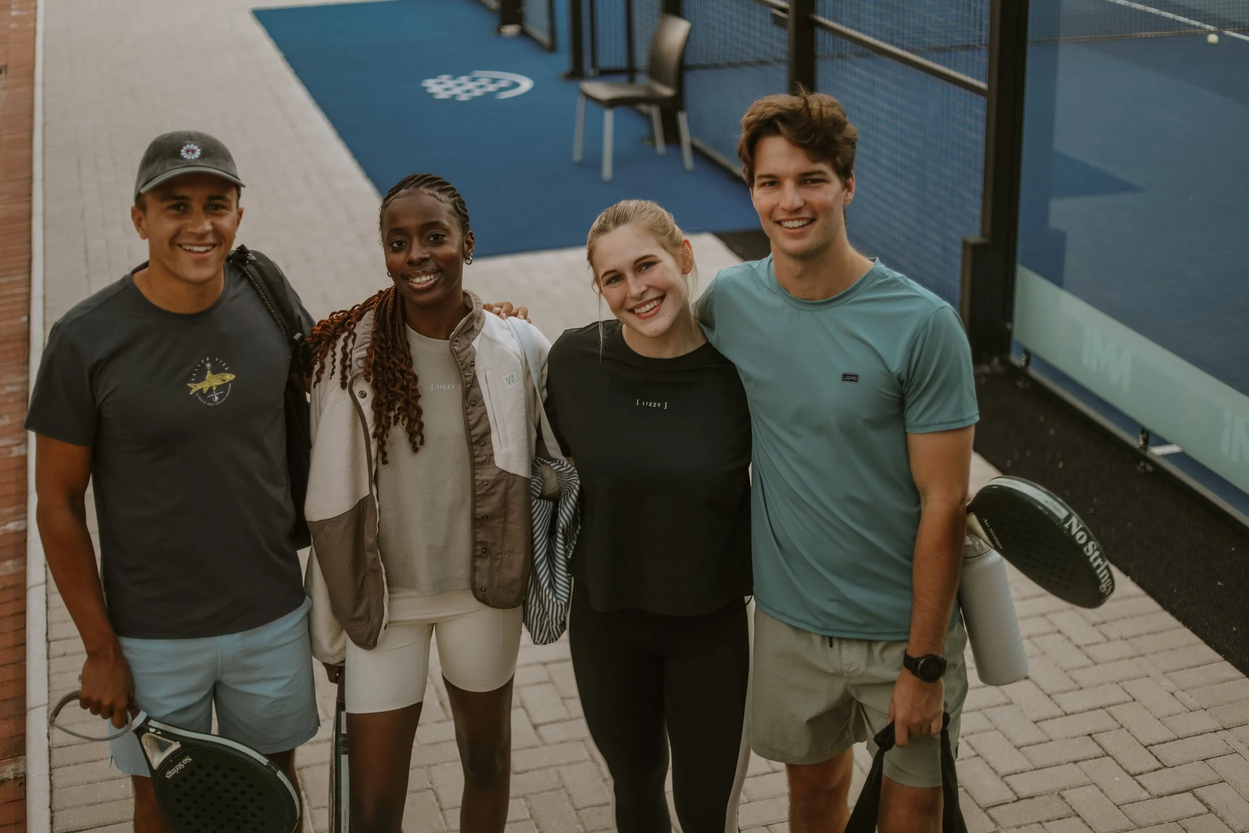 A group of four smiling young adults, two men and two women, standing outside near a tennis or paddle court, holding paddles or rackets, likely after playing a game.