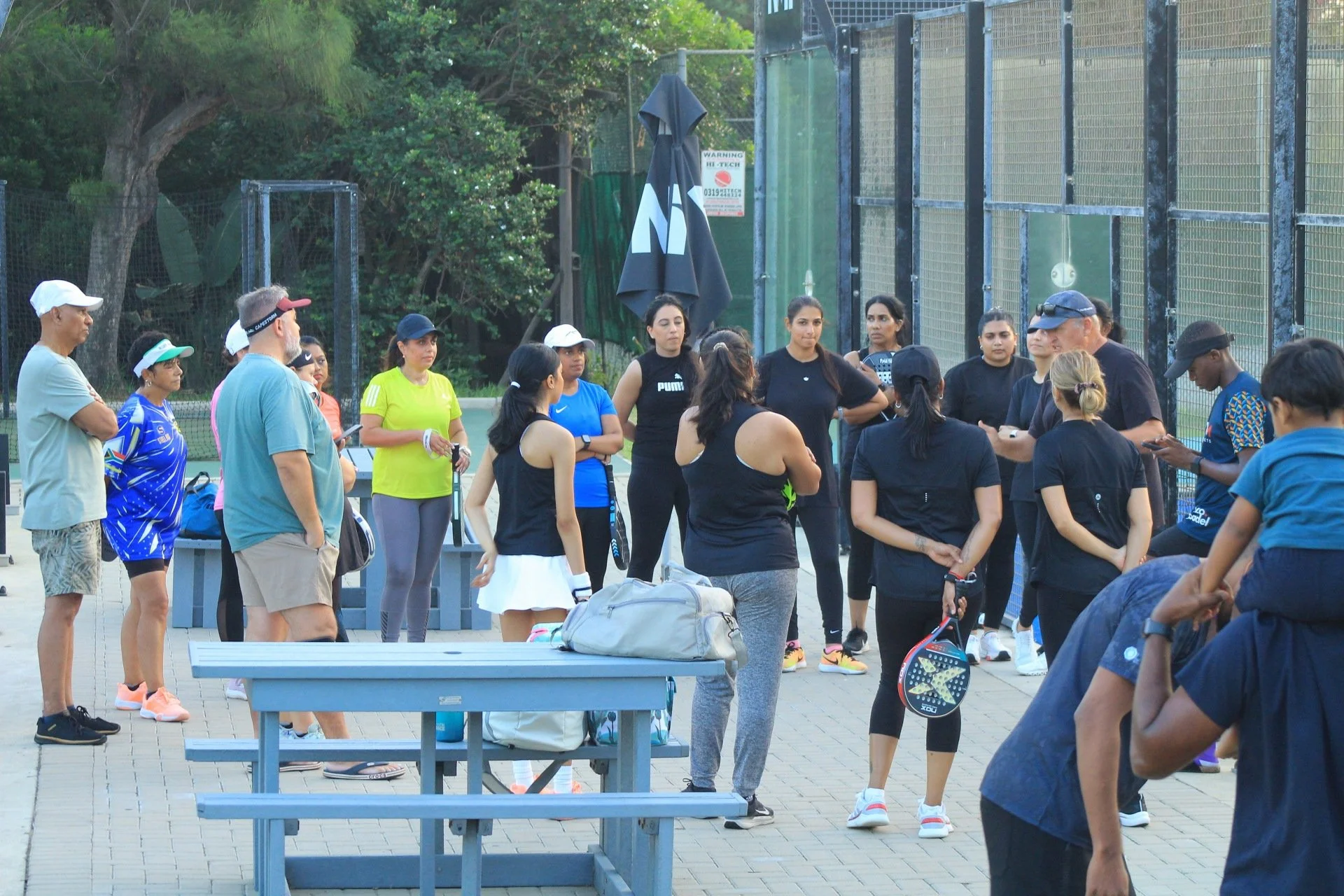 A group of mostly women and a few men gathered on an outdoor tennis court, some holding tennis rackets, in casual athletic clothing, listening to a man who appears to be giving instructions or a talk. There are bags and personal items on a nearby ben