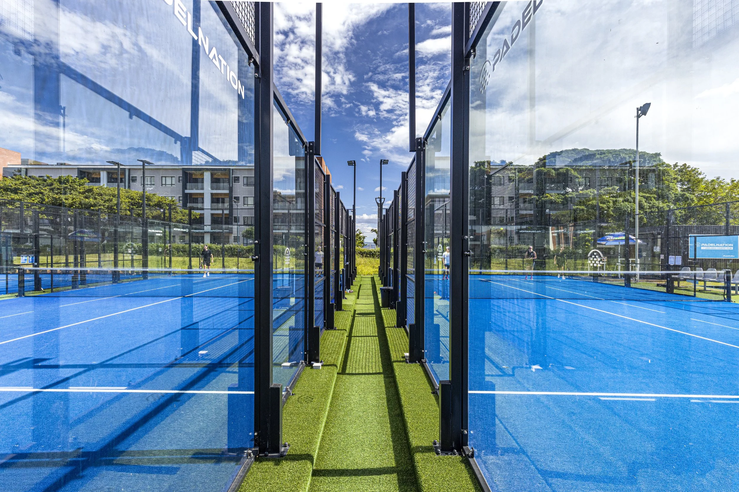 Blue padel courts with black metal and glass enclosures, surrounded by residential buildings and green trees, under a partly cloudy sky.