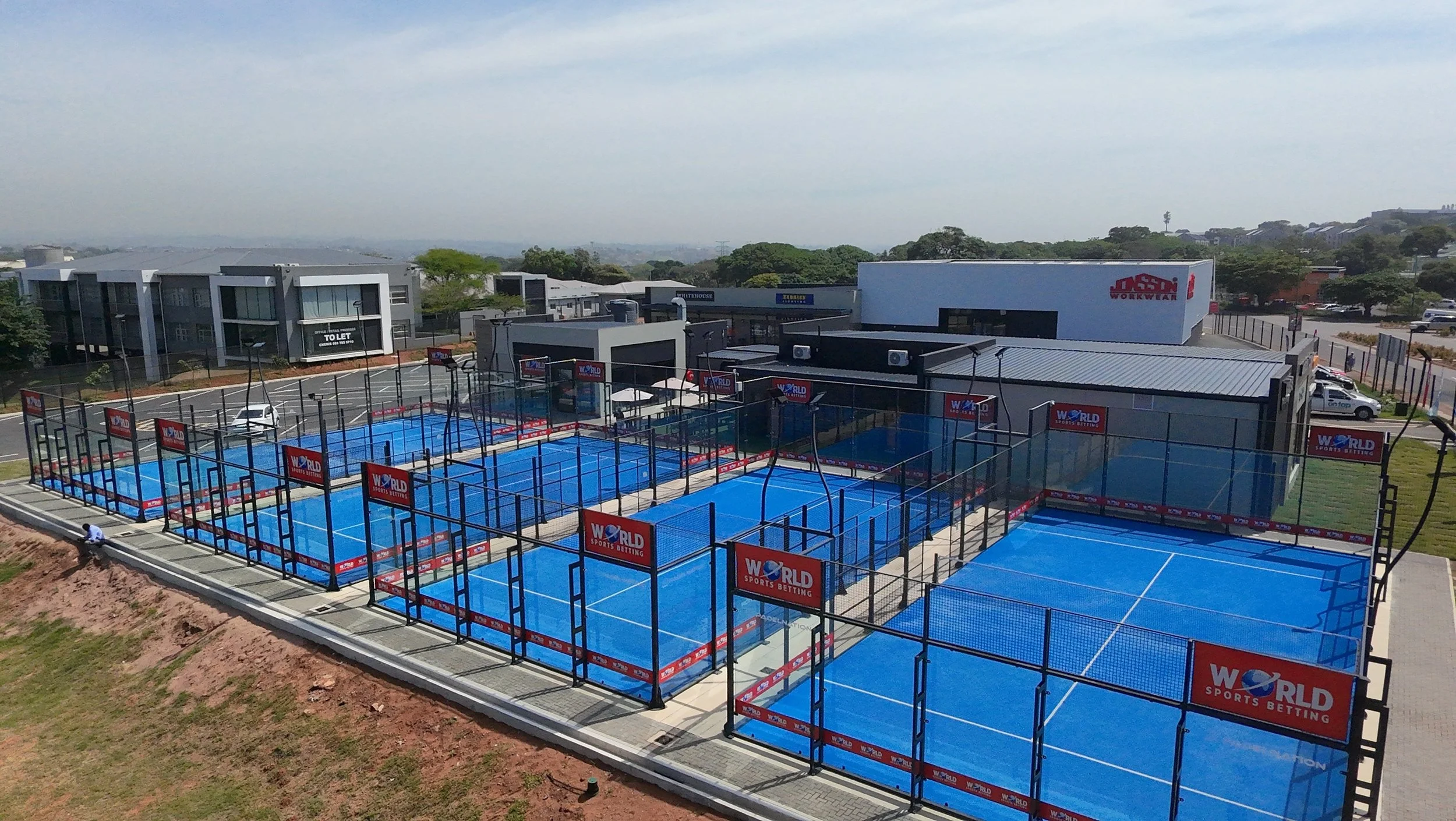Multiple outdoor padel courts with blue surfaces, enclosed by black fences with advertising banners for W-World Sports Betting. Buildings and trees are visible in the background, situated in Amanzimtoti.