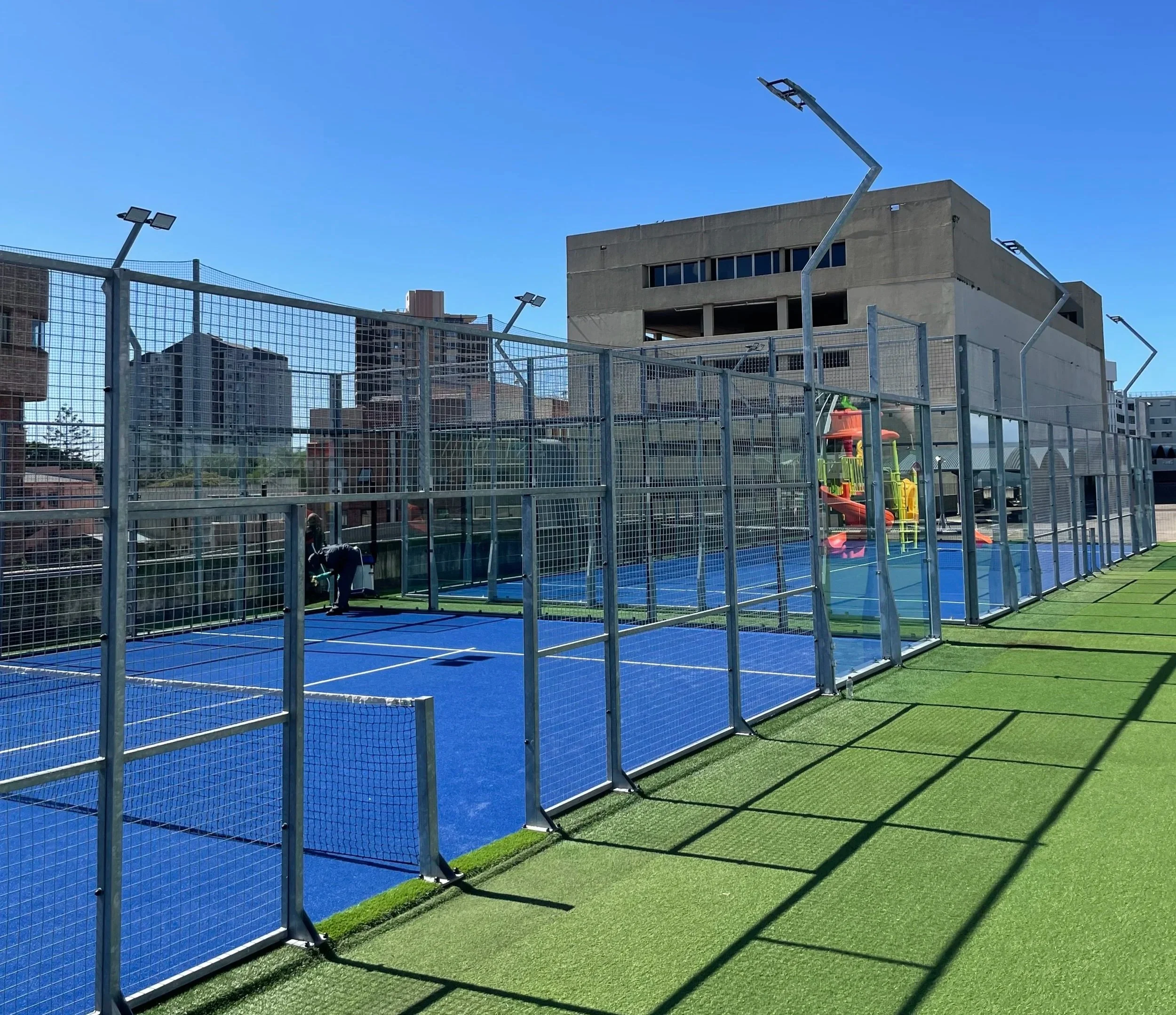 A fenced playground area with blue padel courts and a colorful play structure in the background on a sunny day with a clear blue sky in an urban setting.