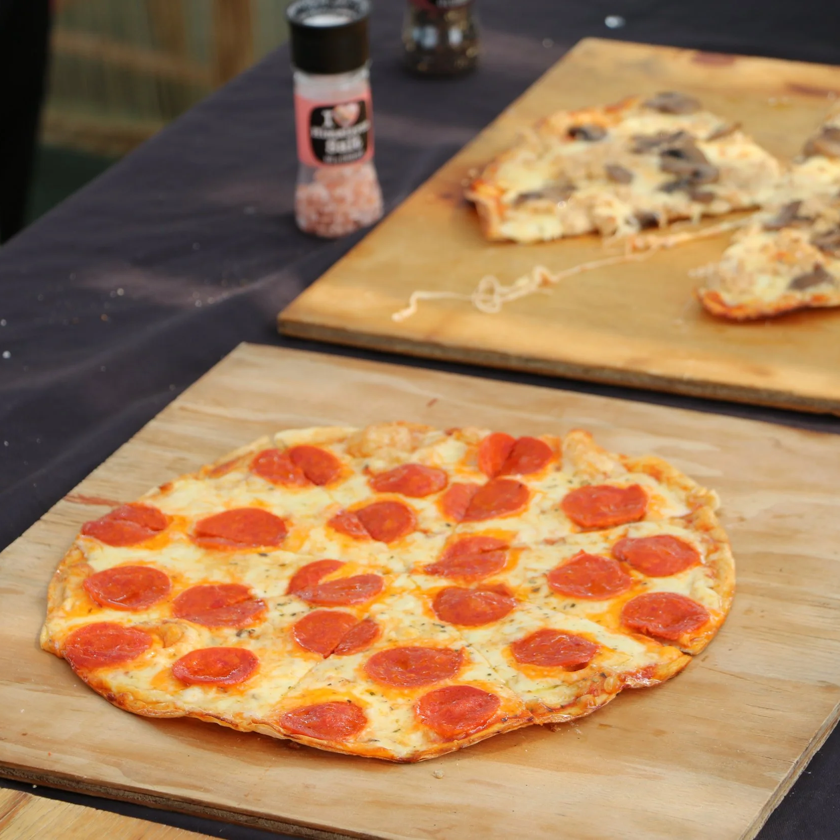 Pepperoni pizza on a wooden paddle on a black table, with a second pizza and salt and pepper shakers in the background.