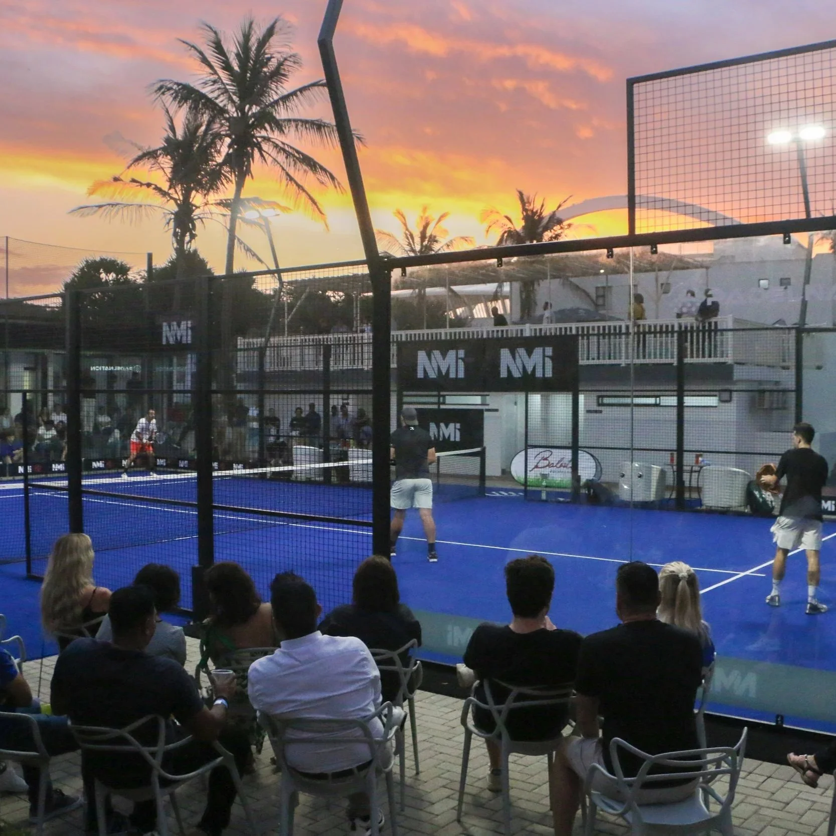 People watching a nighttime outdoor padel match on a blue court, with a sunset sky and palm trees in the background.