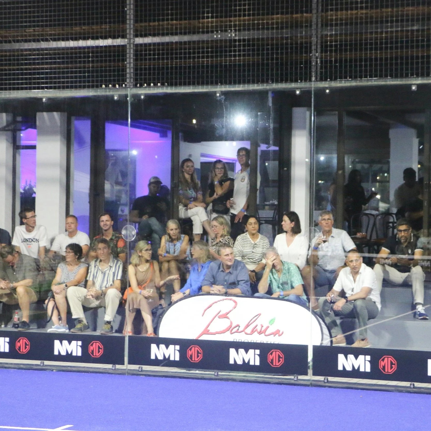 A group of people sitting and standing behind a padel exhibition, with some watching attentively and others talking. The background has a modern interior with purple lighting and an NMI logo on the glass barrier.