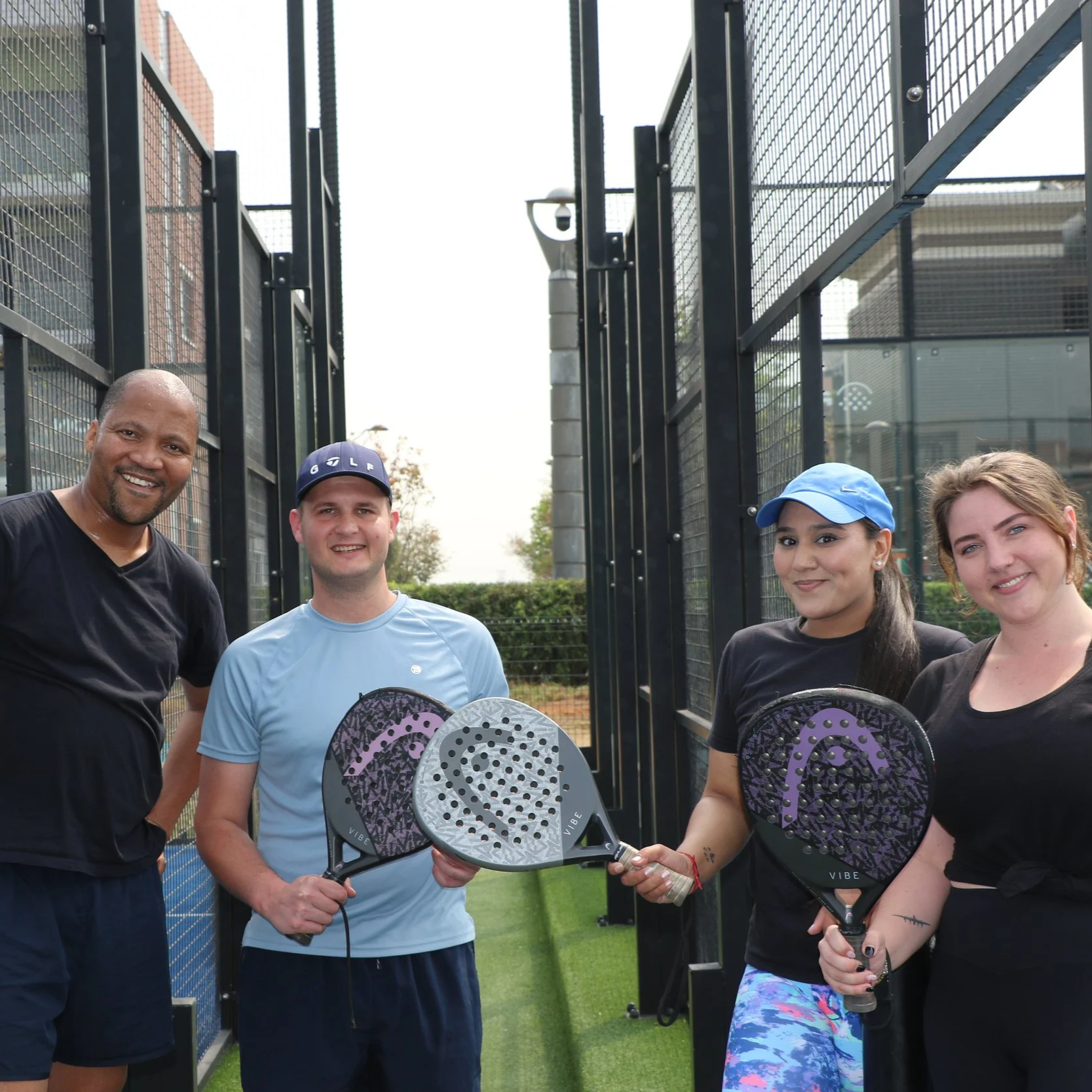 Four people standing outdoors on a padel court, holding paddle rackets and smiling.