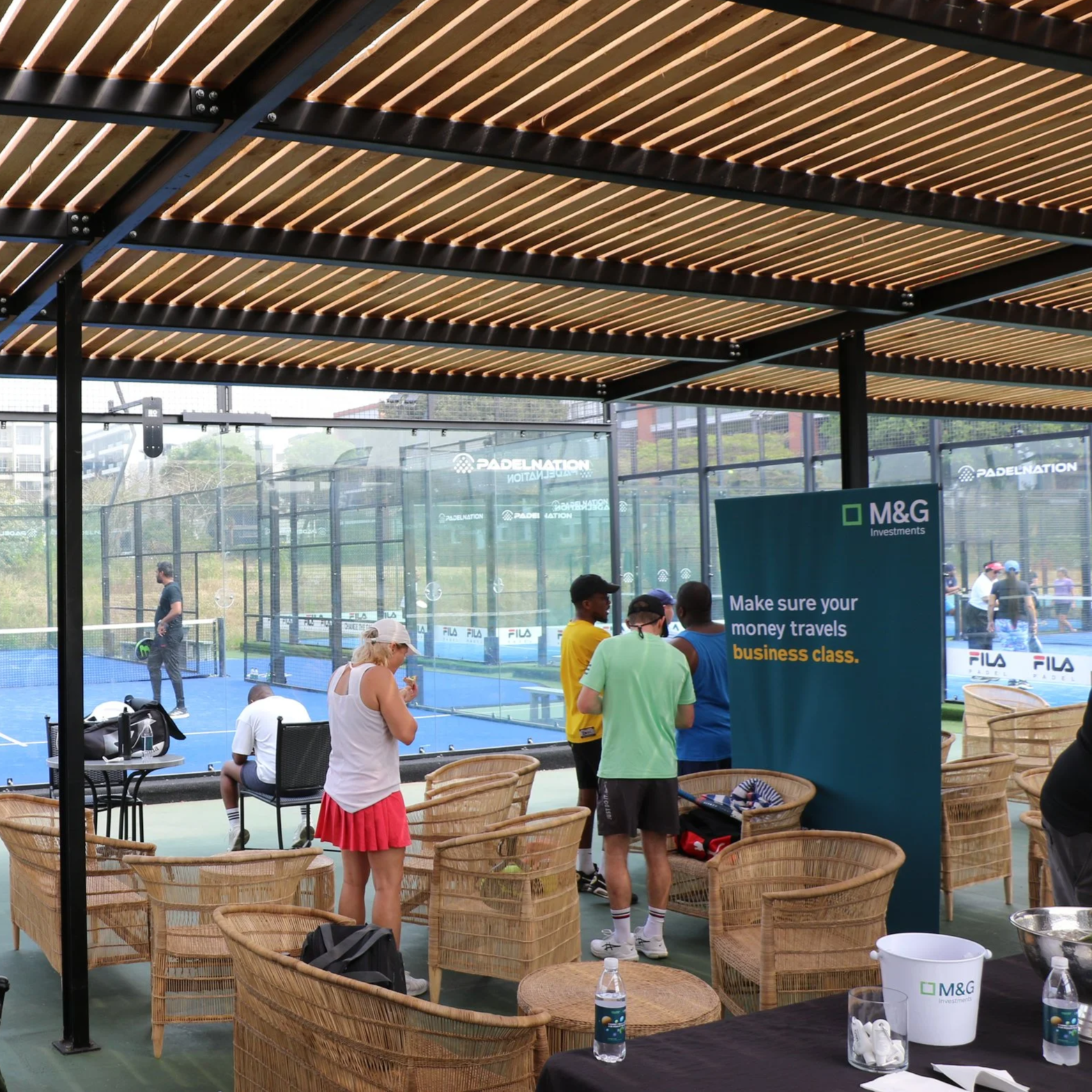 People playing padel  in a court with glass walls, under a wooden slatted ceiling, while others are gathered around a table with promotional banners at Umhlanga Country Club.