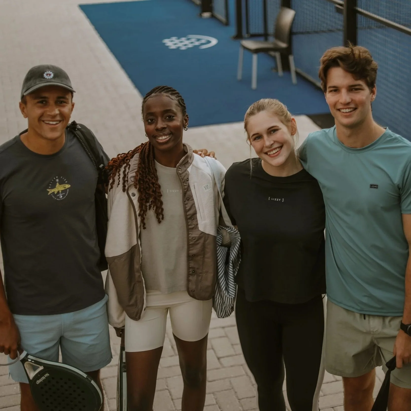 Four young adults standing together outdoors, smiling, with a blue padel court in the background.
