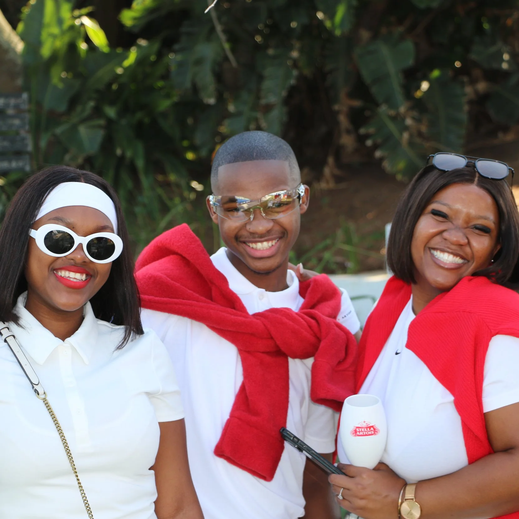Three smiling women and one smiling man at a padel event at Durban Country Club, dressed casually with sunglasses, holding a Stella Artois glass, with greenery in the background.