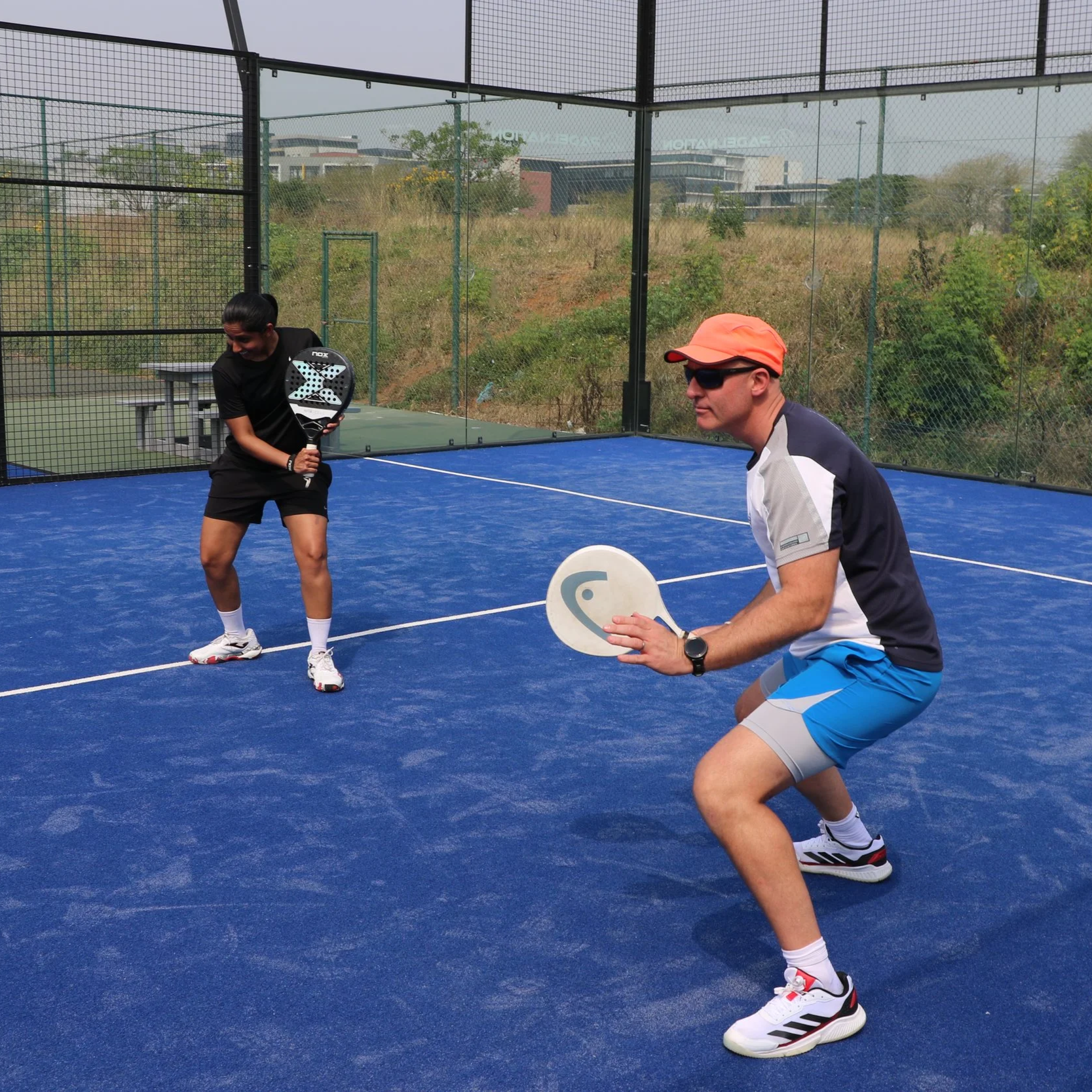 Two people having fun playing padel on an outdoor court with a blue surface, surrounded by a chain-link fence, with trees and buildings in the background.