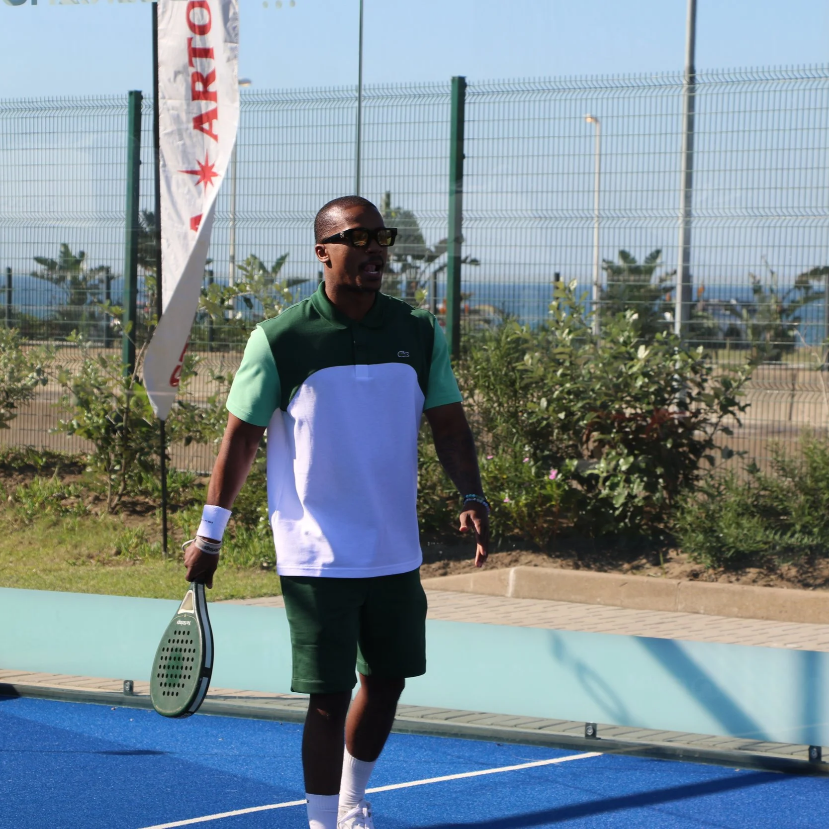 A man standing on a padel court holding a padel racquet, wearing sunglasses, a color-blocked polo shirt, shorts, and white sneakers in an outdoor setting with a fence, plants, and a clear sky.