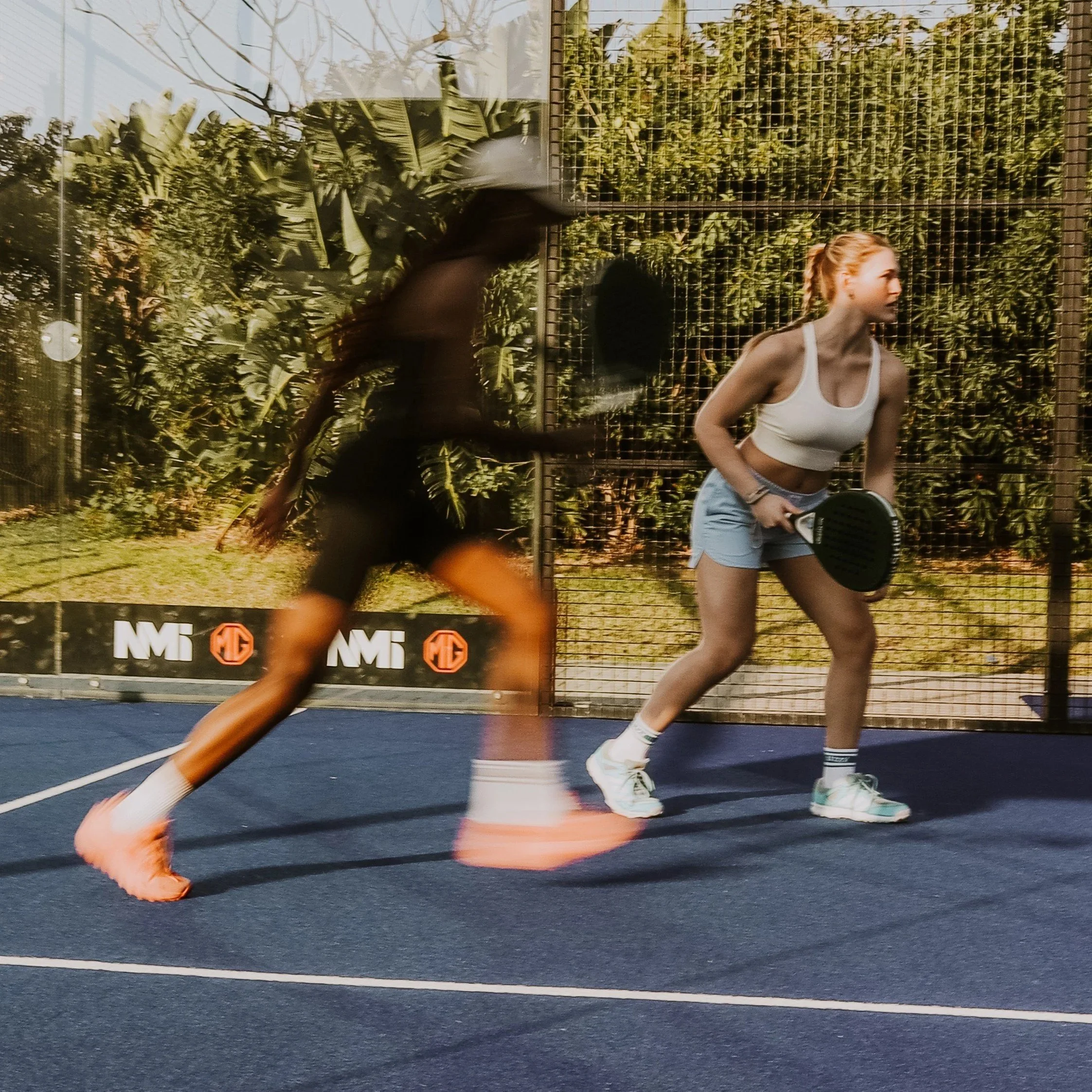 Two women playing padel on an outdoor court, one preparing to hit the ball while the other is running past.