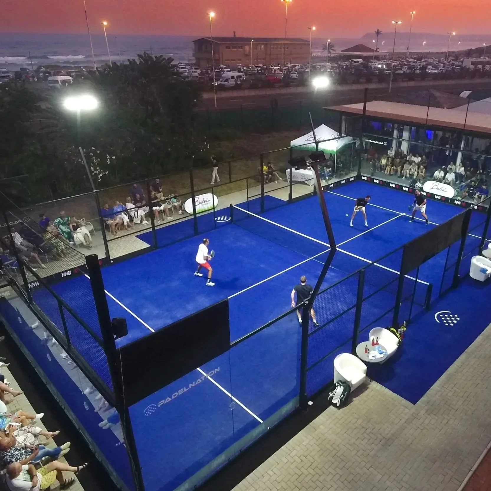 A blue padel court at sunset with four players competing, surrounded by spectators seated on benches and standing outside the fenced court.