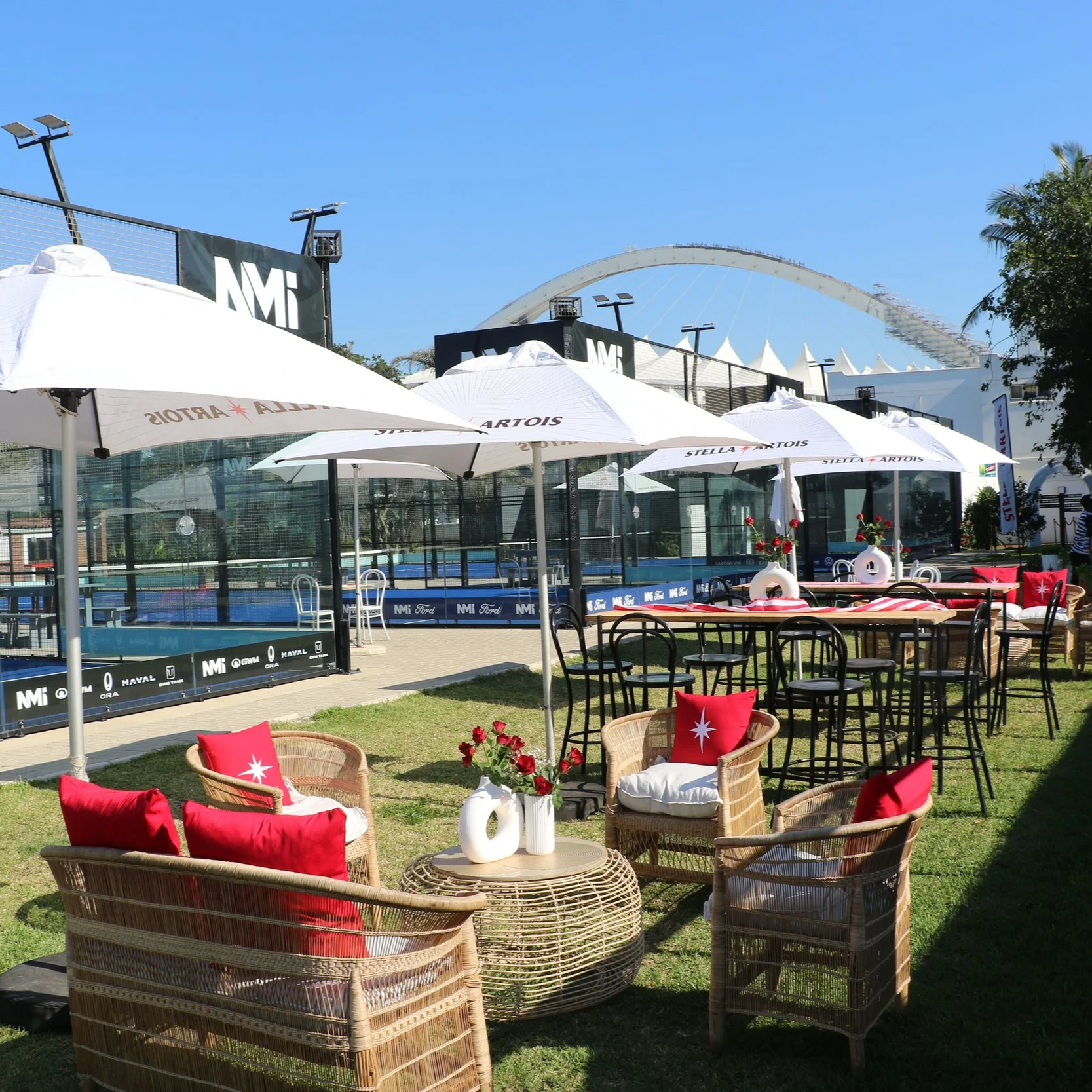 Outdoor seating area with wicker chairs and red pillows around a round wicker table with vases of red flowers, shaded by large white umbrellas, with a padel court and Moses Mabida Stadium in the background.