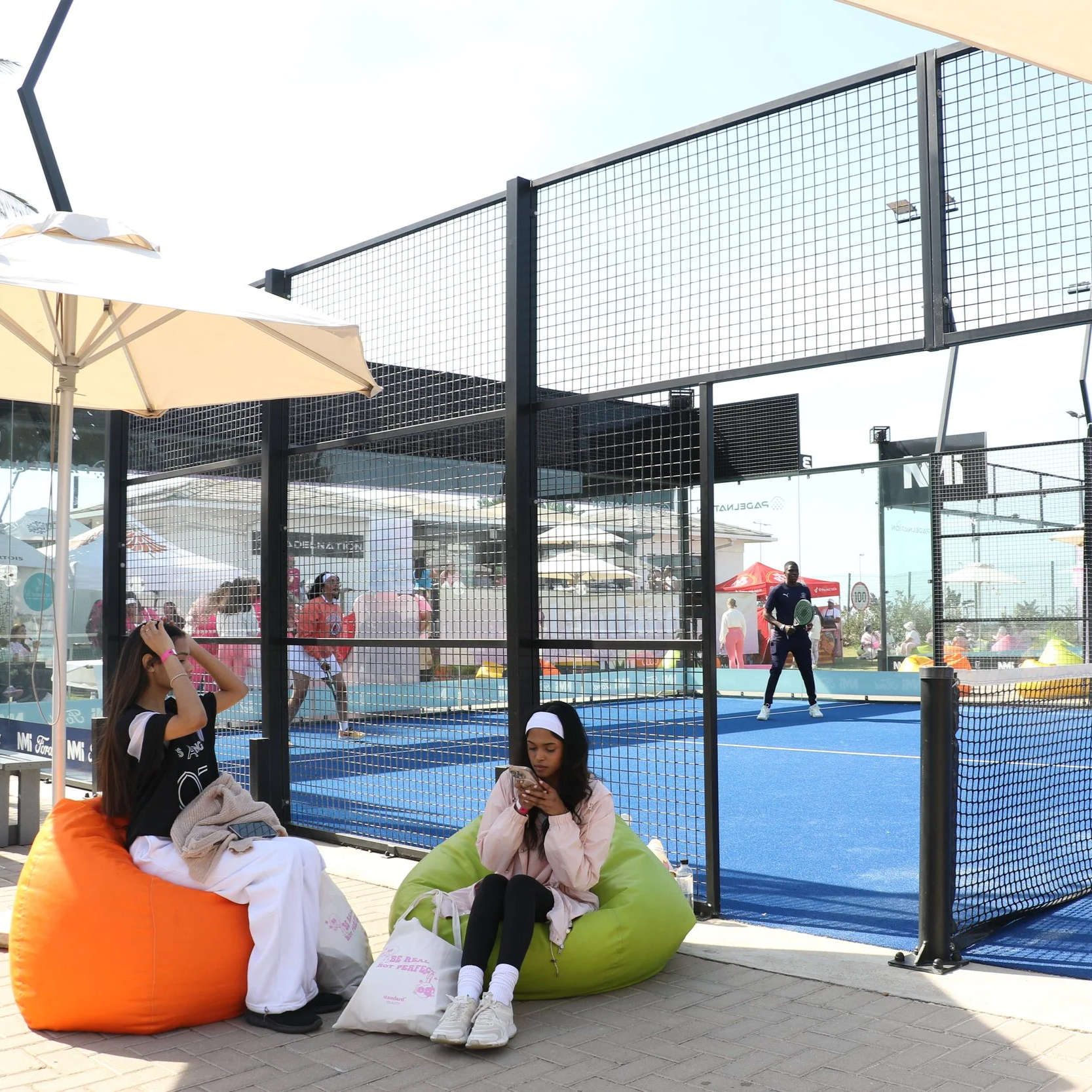 Two young women sitting on bean bags outside a padel court, with one looking at her phone and the other adjusting her hair. The court is enclosed by a black metal fence, with people playing padel inside on blue turf courts.
