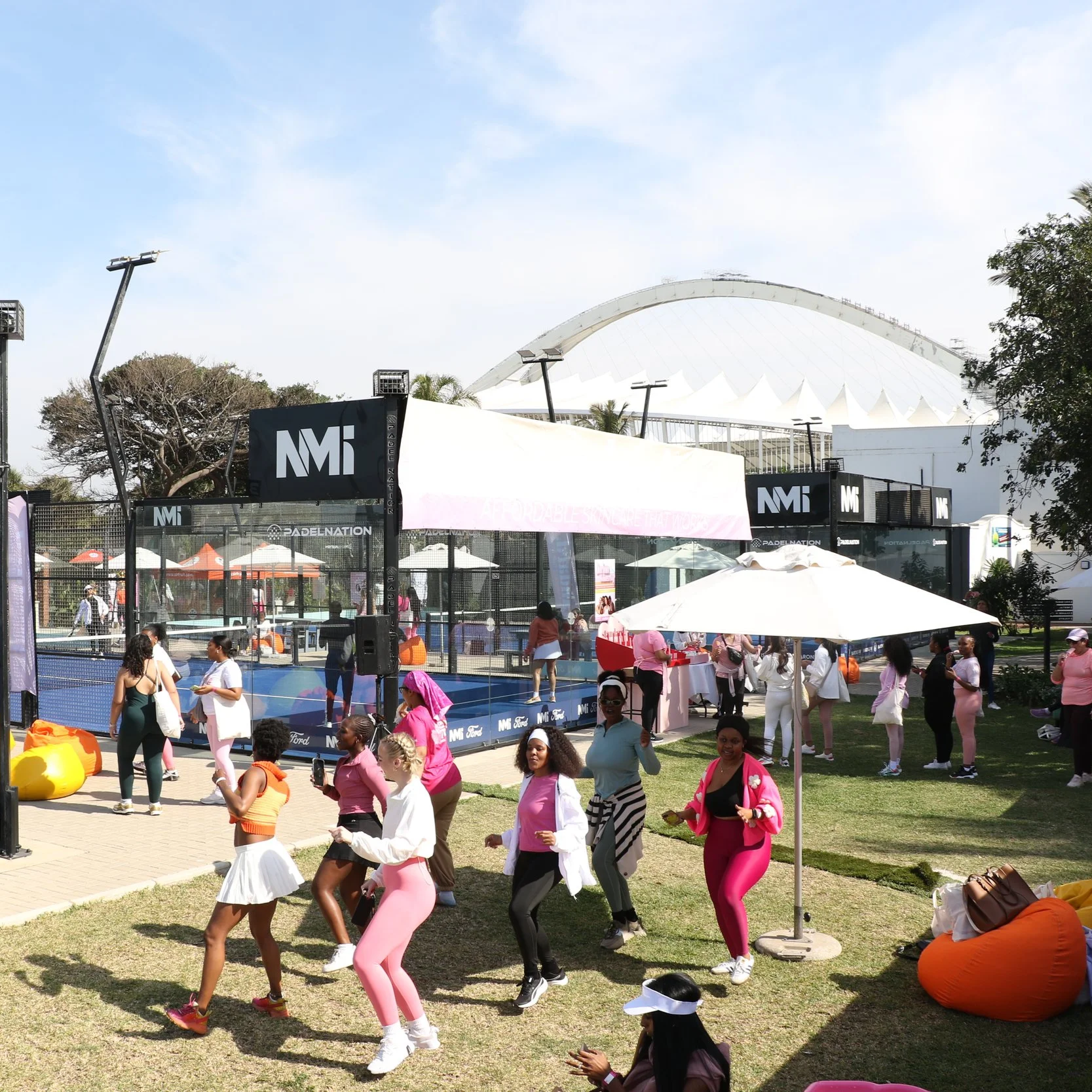 Outdoor event with people playing padel at Durban Country Club, socialising, and dancing under umbrellas and tents with the Moses Mabida Stadium in the background.