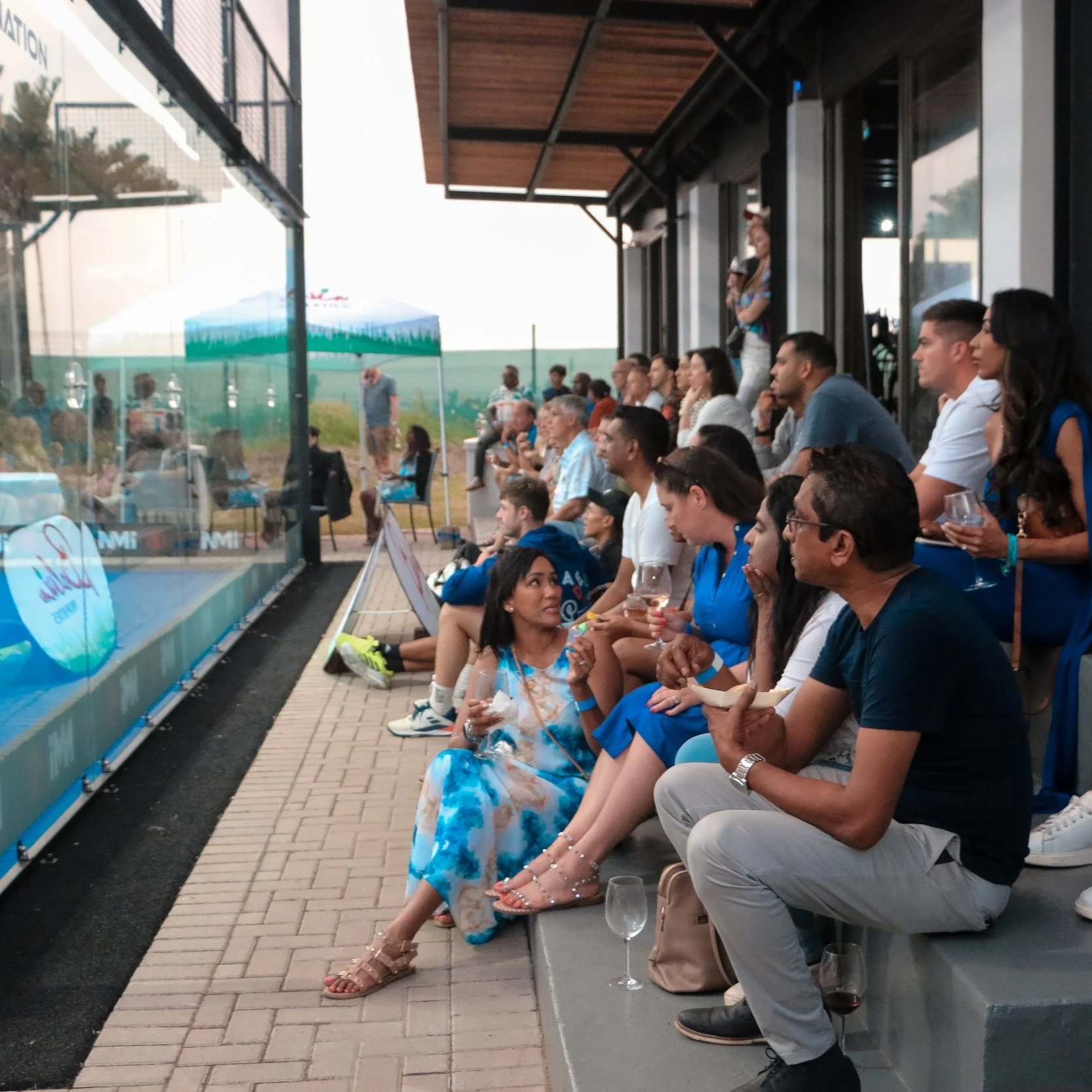 People seated on outdoor tiered seating watching a padel match, some holding drinks, under a covered patio at Durban Country Club.