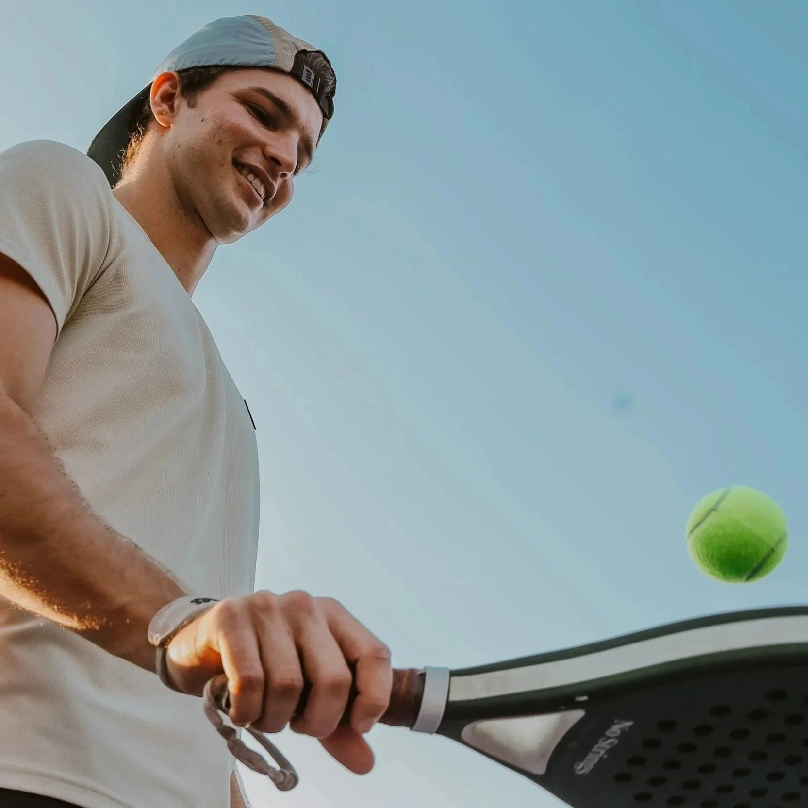 A young man wearing a backward baseball cap and beige shirt smiling while holding a padel racket, with a padel ball approaching.