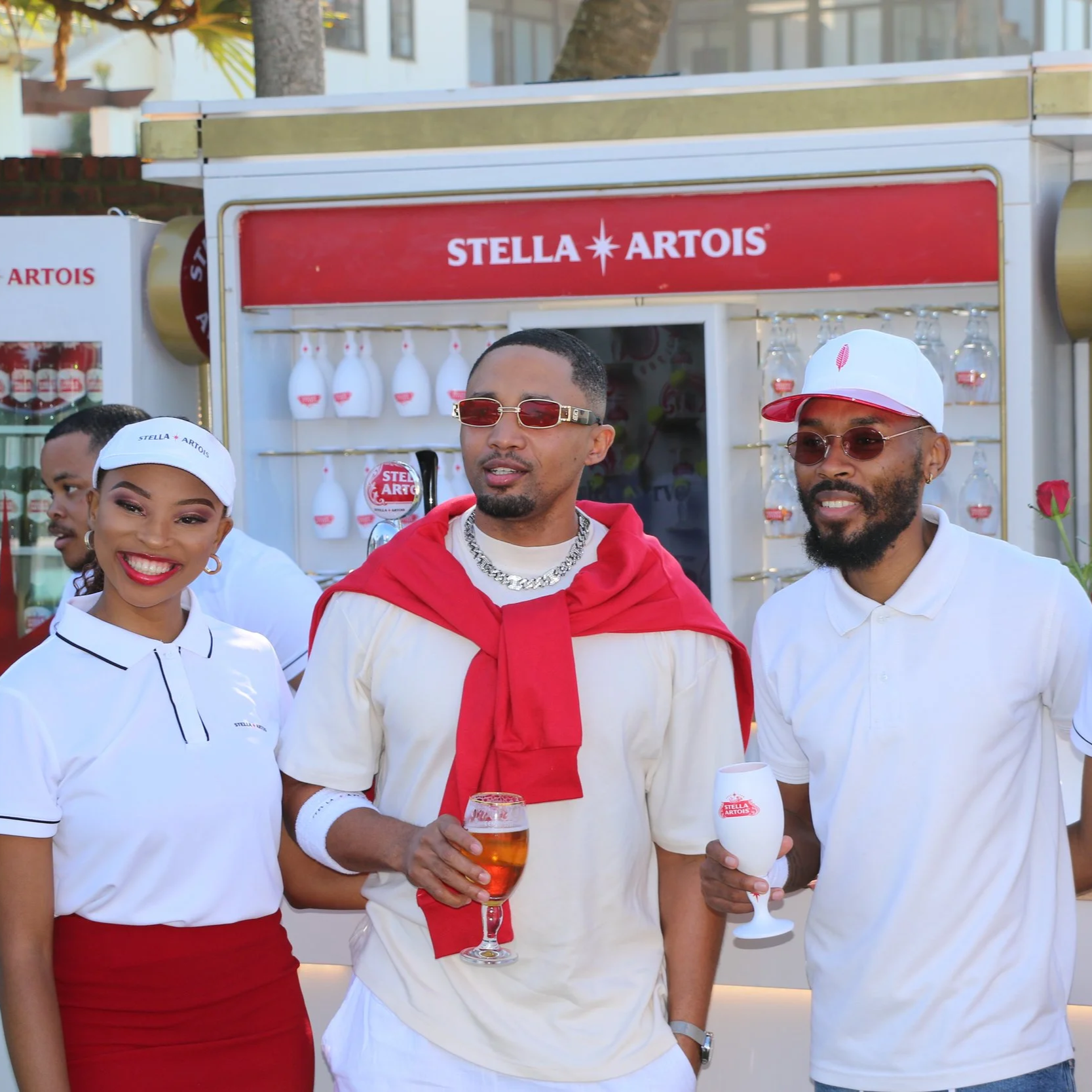 Group of young adults at a Stella Artois event, standing in front of a Stella Artois branded booth, holding drinks, smiling, dressed casually with sunglasses.