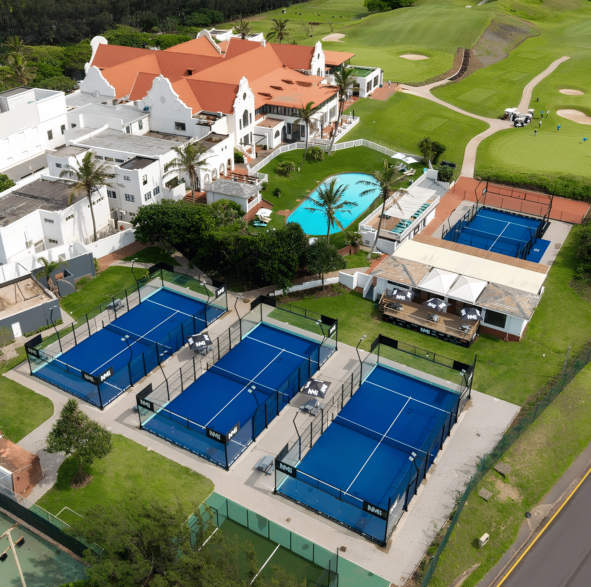 Aerial view of a luxury golf resort with three padel tennis courts, a swimming pool with palm trees, a small building with outdoor seating, and a golf course with sand traps and greens.