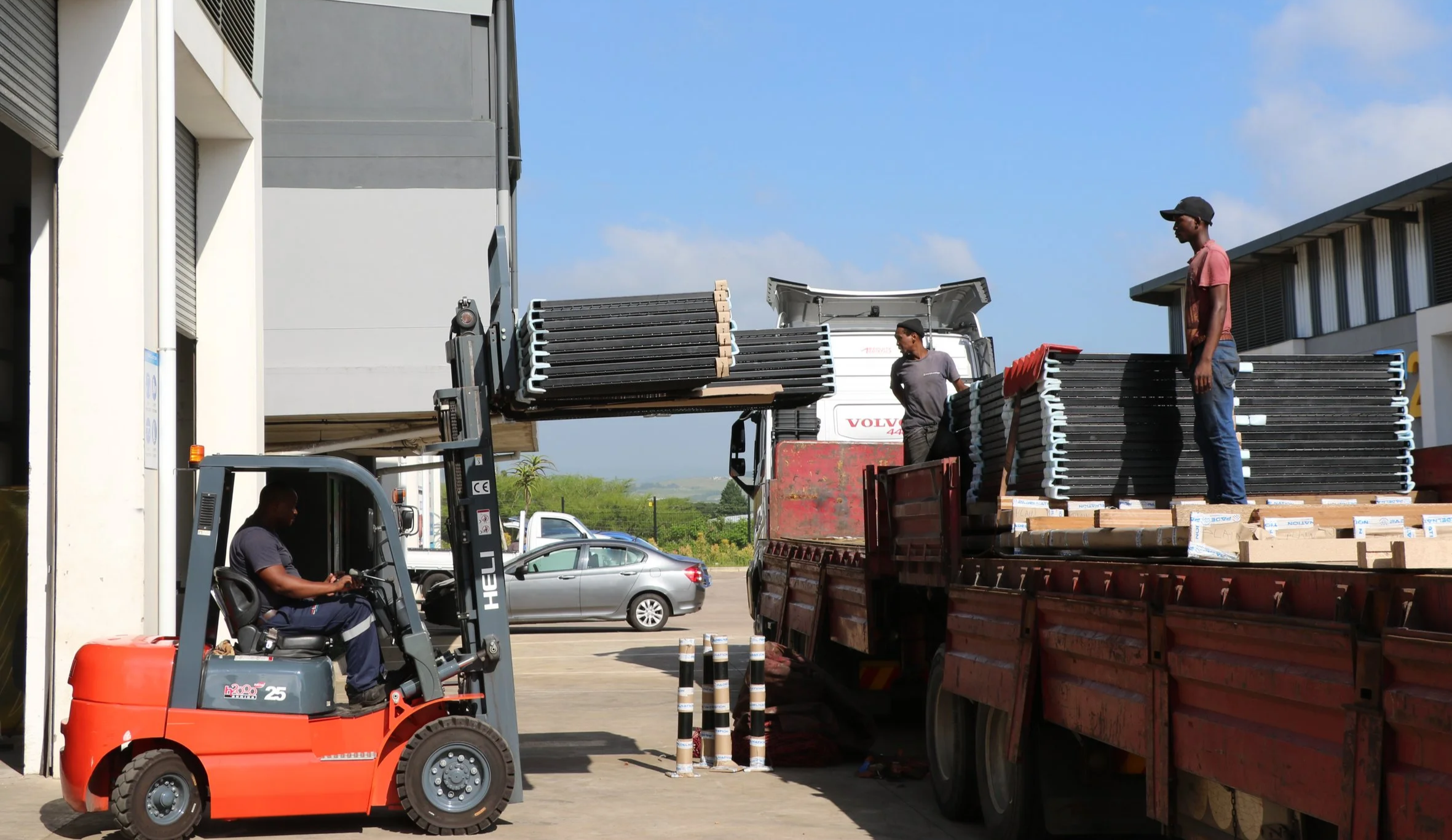 Workers unloading and stacking chairs from a truck using a forklift outside a building on a sunny day.