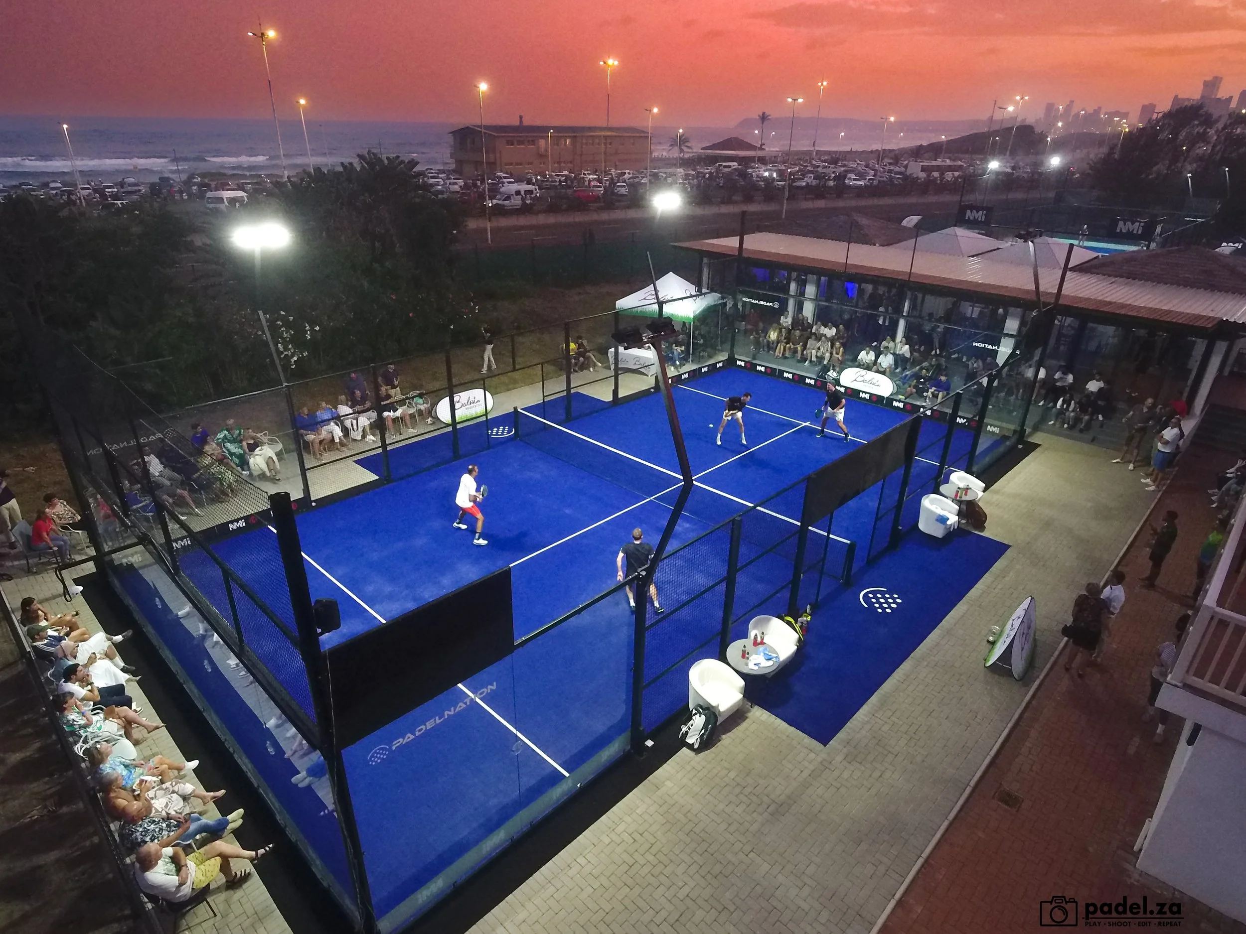 An outdoor paddle tennis court at sunset with players actively engaged, surrounded by spectators seated outside and a scenic ocean view in the background.