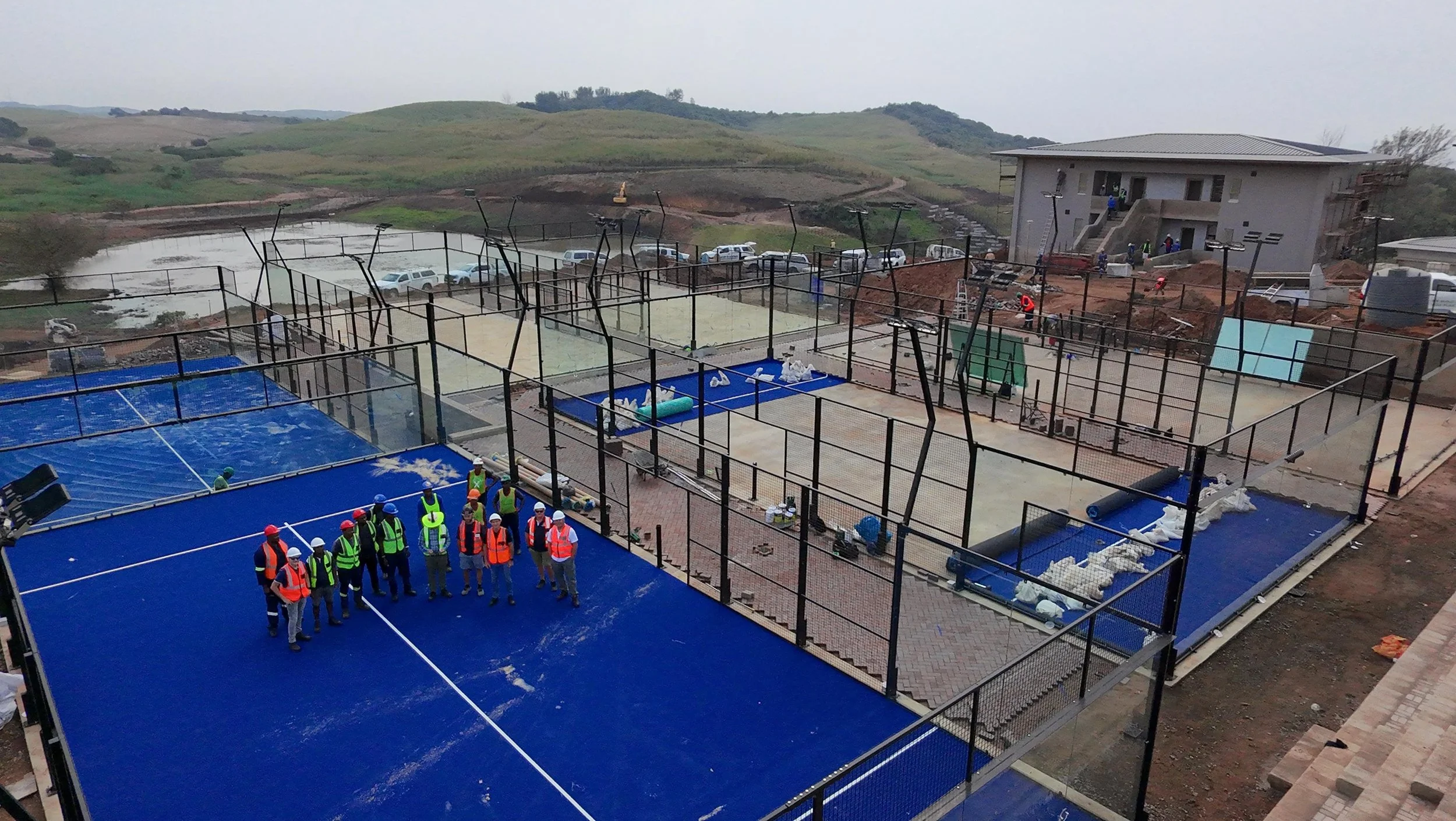 Construction workers in safety gear inspecting a sports complex with multiple enclosed courts under construction, surrounded by hilly landscape.