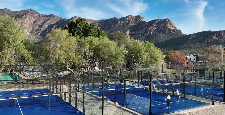 Multiple tennis courts with blue surfaces and netting, surrounded by trees and mountains in the background, under a partly cloudy sky.