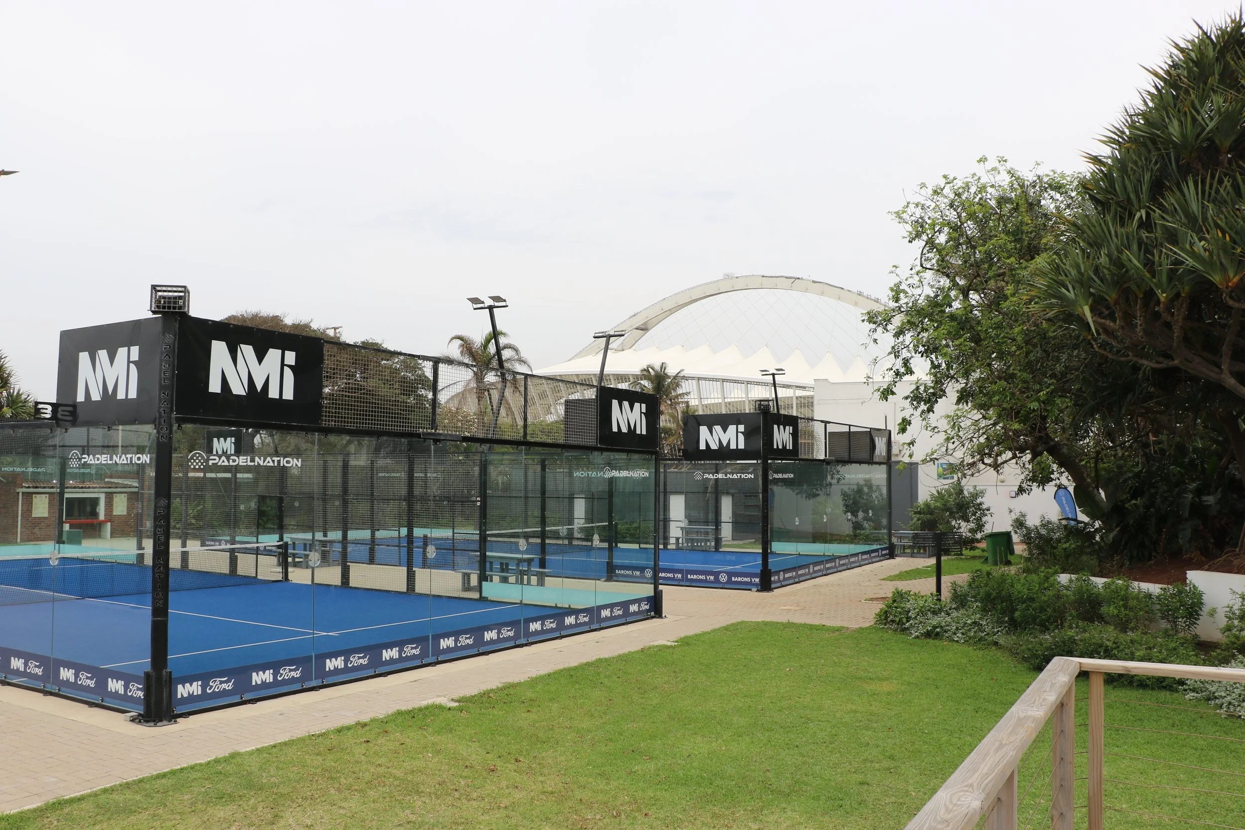Outdoor padel tennis courts with blue surfaces and surrounding fencing, branded with logos, located in a well-maintained grassy area with trees and a pathway.