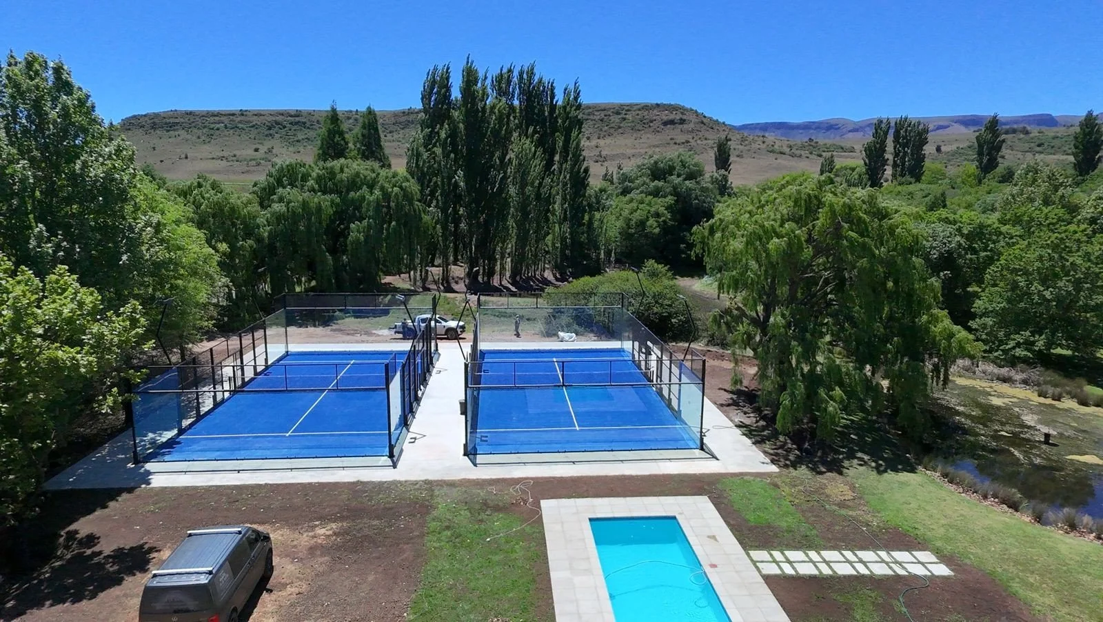 A backyard with two covered tennis courts, a swimming pool, and multiple trees in a rural area with hills in the background.