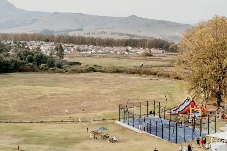 Children playing on a playground slide near a fenced tennis or basketball court, with people sitting on benches and a group gathered nearby, in a park-like setting with open fields, trees, and hills in the background.