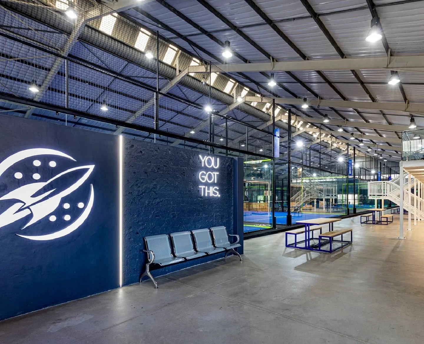 Interior of an indoor trampoline park with seating area, blue and black decor, and neon sign that reads 'You got this.'