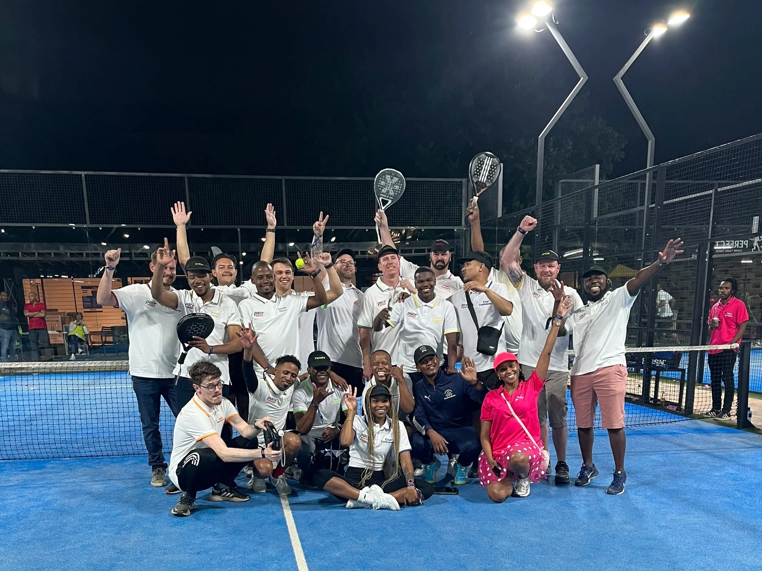 Group of tennis players and supporters celebrating on a blue tennis court at night, holding rackets and raising their hands.