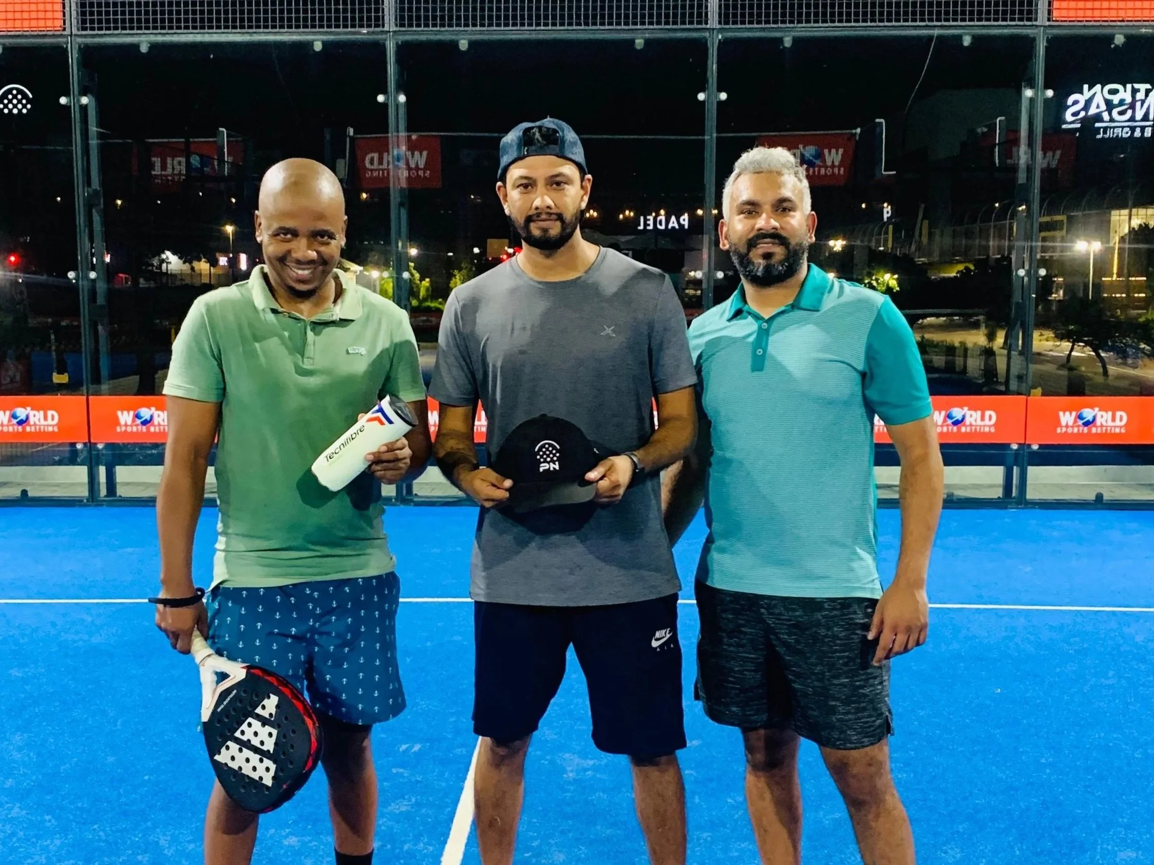 Three men standing on a blue tennis court at night, holding tennis rackets and a hat, with glass barriers and city lights in the background.
