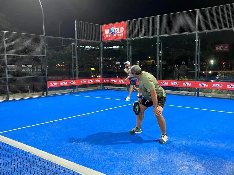 Two men playing pickleball on a blue court at night, with a fence around it and signage that reads 'World Sports Betting'.