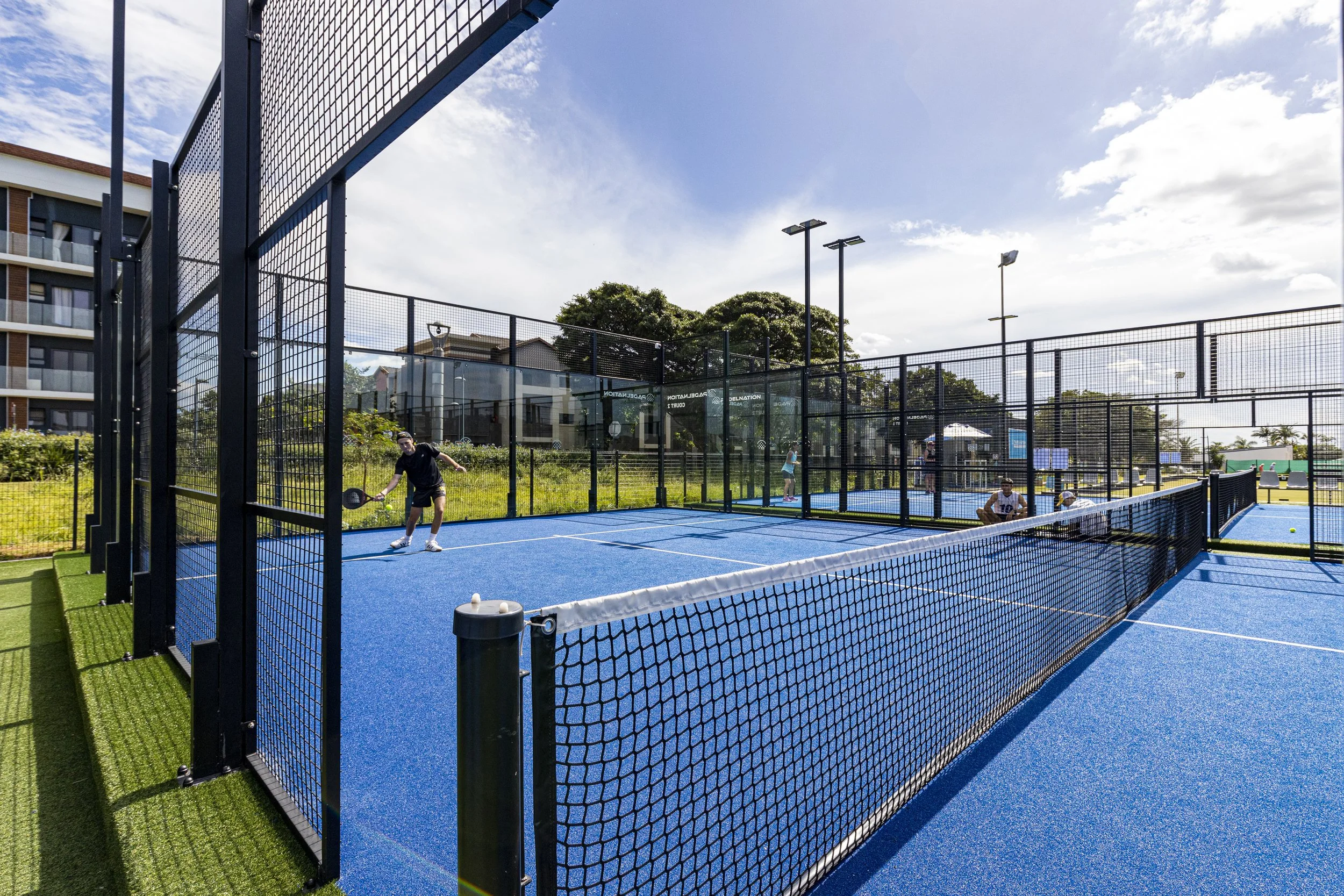 A tennis court with blue surface surrounded by black fencing. There are people playing tennis, one of them is serving. The court is outdoors with a partly cloudy sky and trees in the background.
