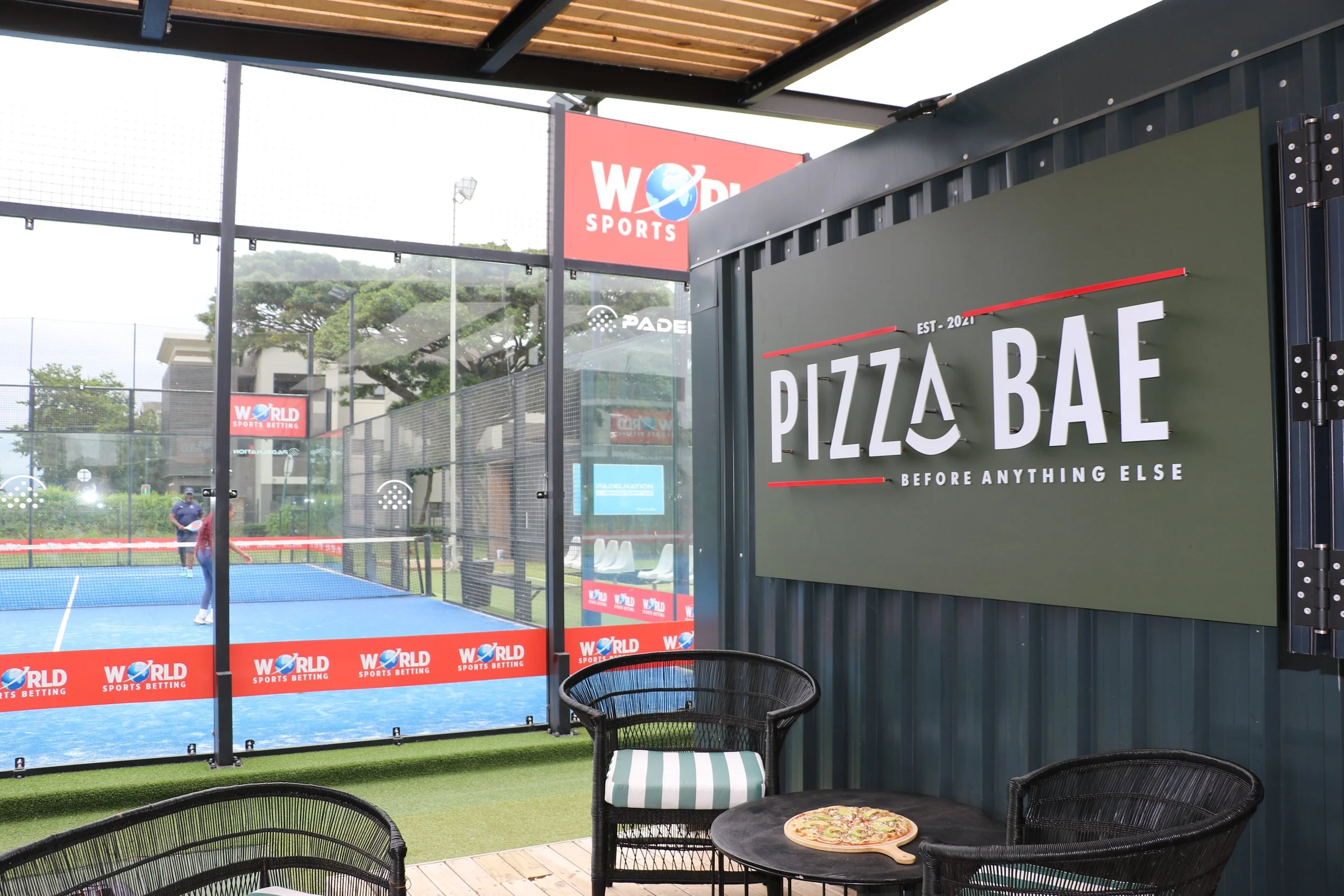 Outdoor padel tennis court viewed from a covered seating area with a pizza on a small table in the foreground.
