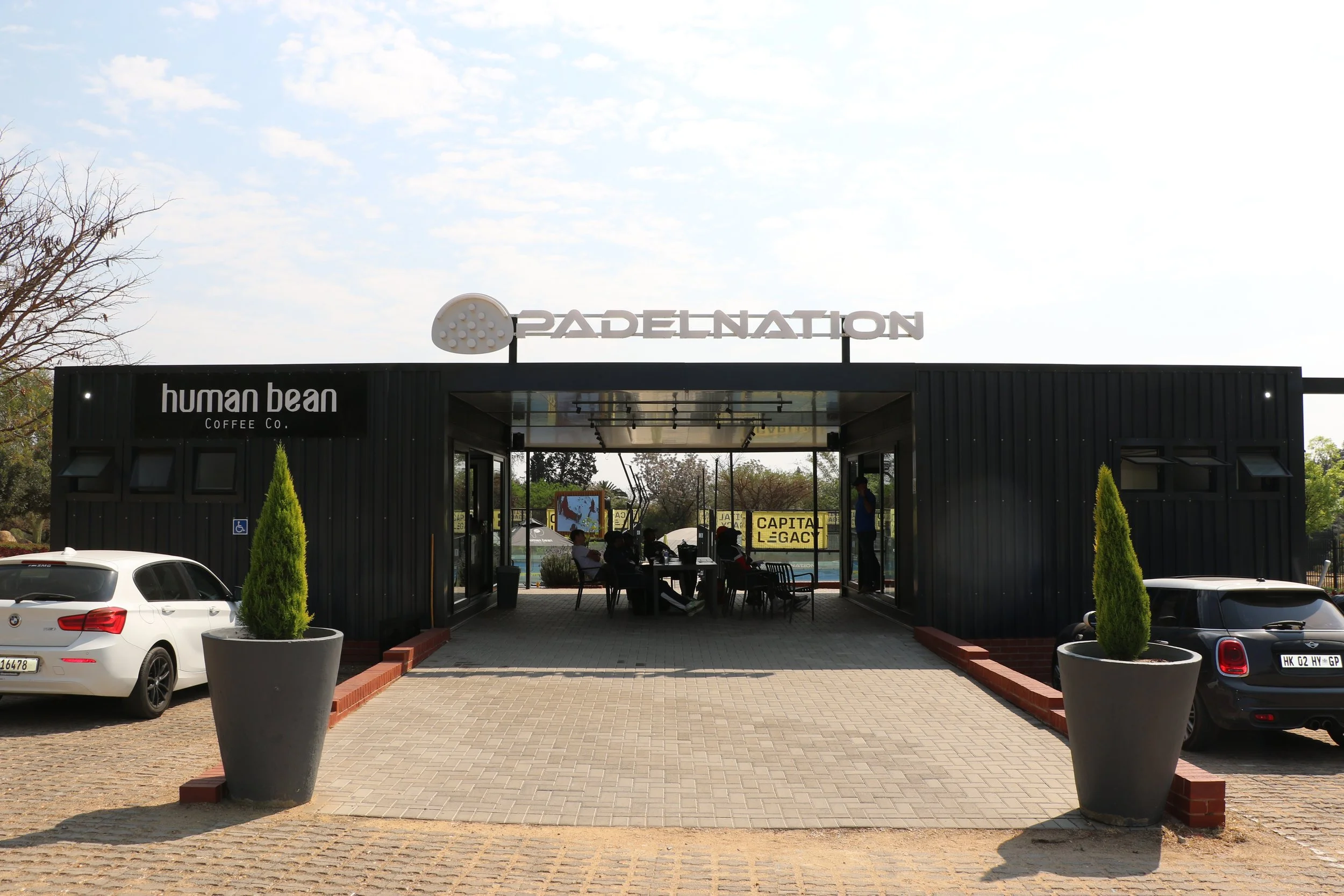 Outdoor scene of a padel tennis court entrance with signs for 'PadelNation' and 'Human Bean Coffee Co.'; cars parked in front; potted plants and trees around; people sitting and standing inside the entrance under a clear sky.