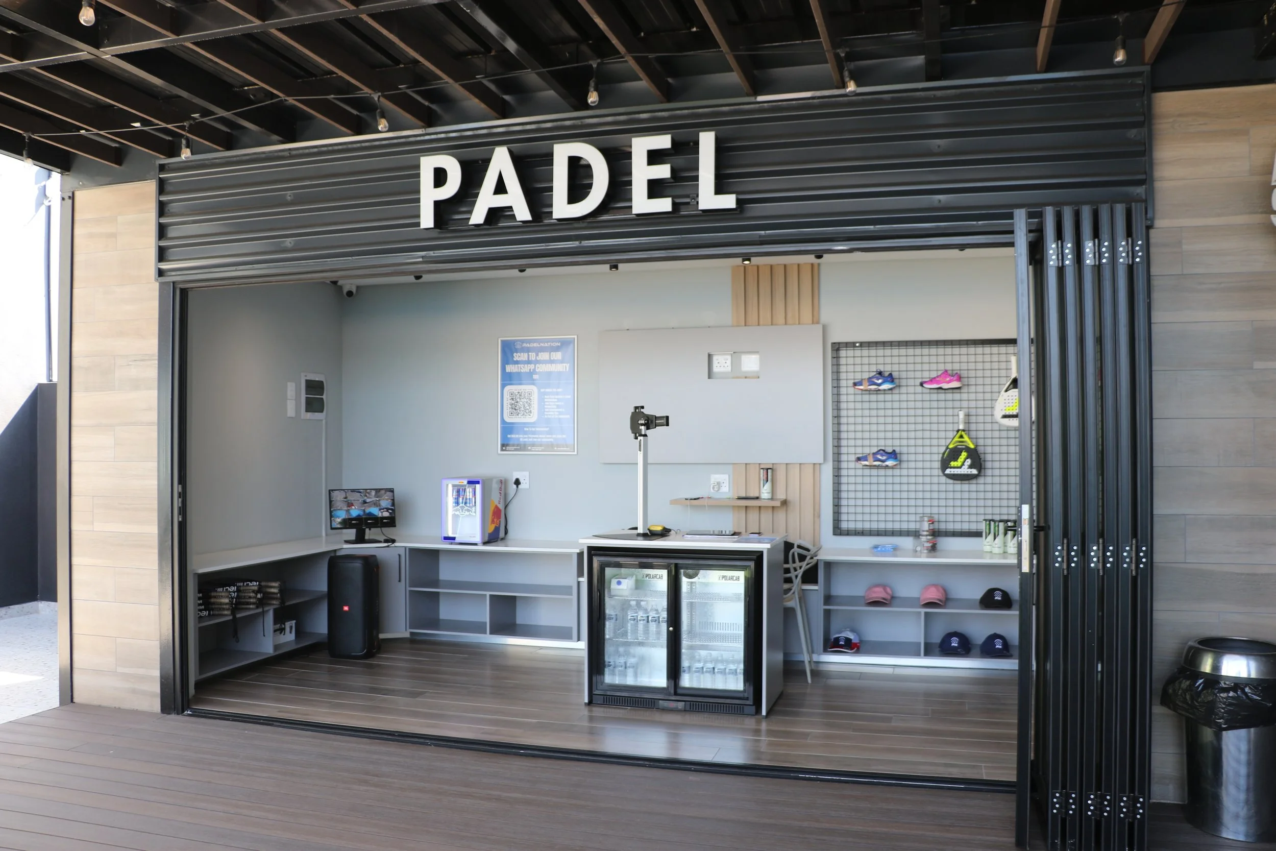 A modern indoor padel court entrance with a sign reading 'PADEL' in large white letters, featuring a small retail area with shoes, caps, and a refrigerator, and technological equipment including a computer and a camera.